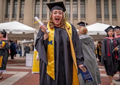 Standing outside Hill Auditorium, a graduate in academic regalia holds up the scroll they received on the Commencement stage with an excited smile.