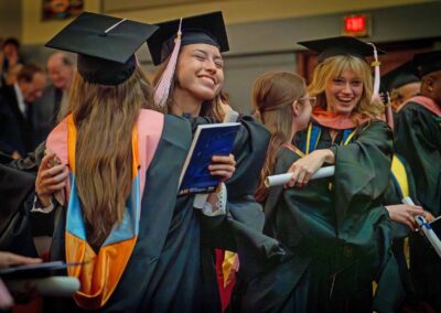 Graduates wearing academic regalia smile and hug inside the house at Hill Auditorium.
