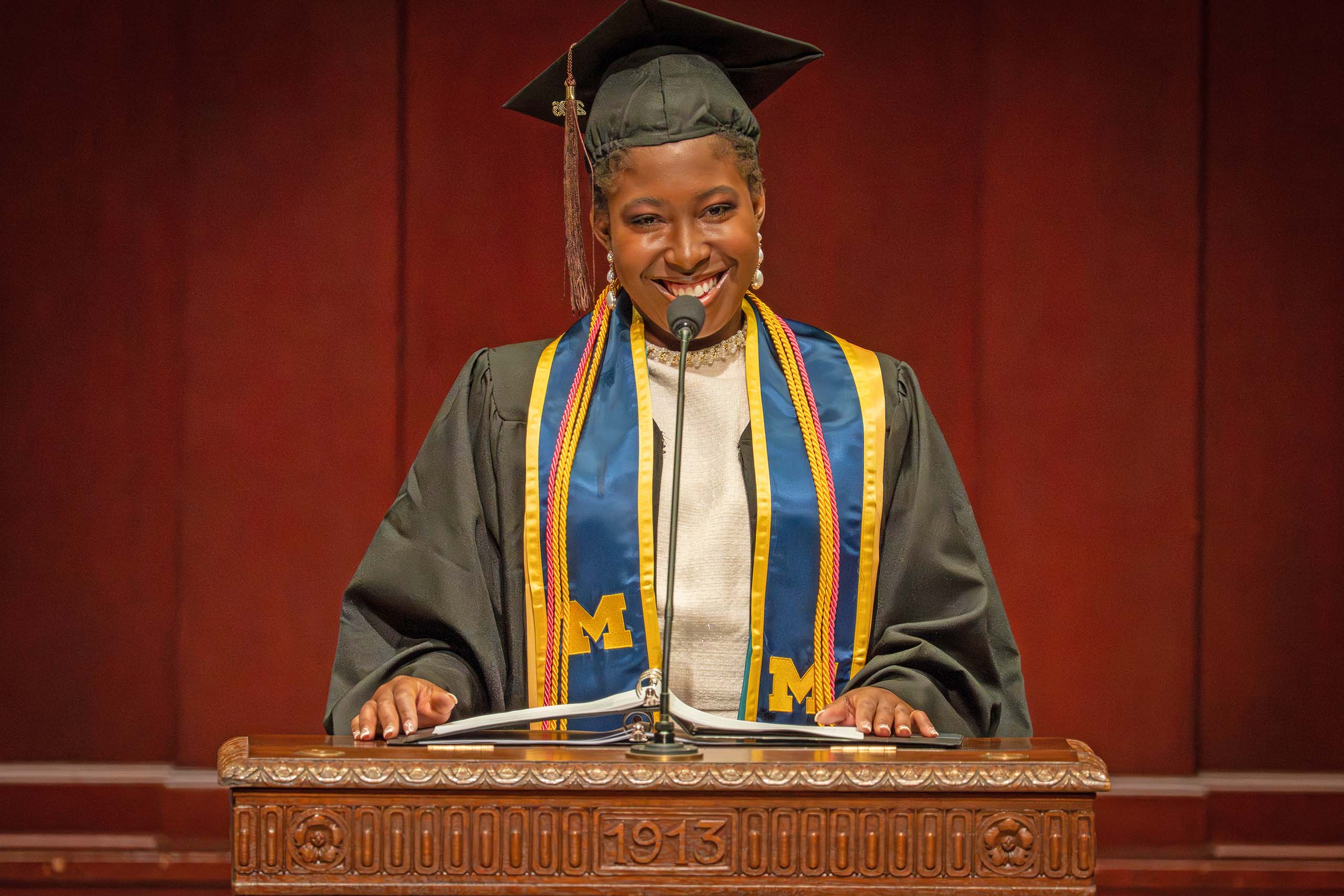 Kristabel Kenta-Bibi speaks at the podium at Hill Auditorium, attired in graduation cap and gown.