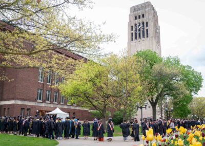 Graduates wearing academic regalia line up outside Hill Auditorium, with Burton Tower in the distance. Spring leaves are growing on trees and a bed of tulips is in bloom.