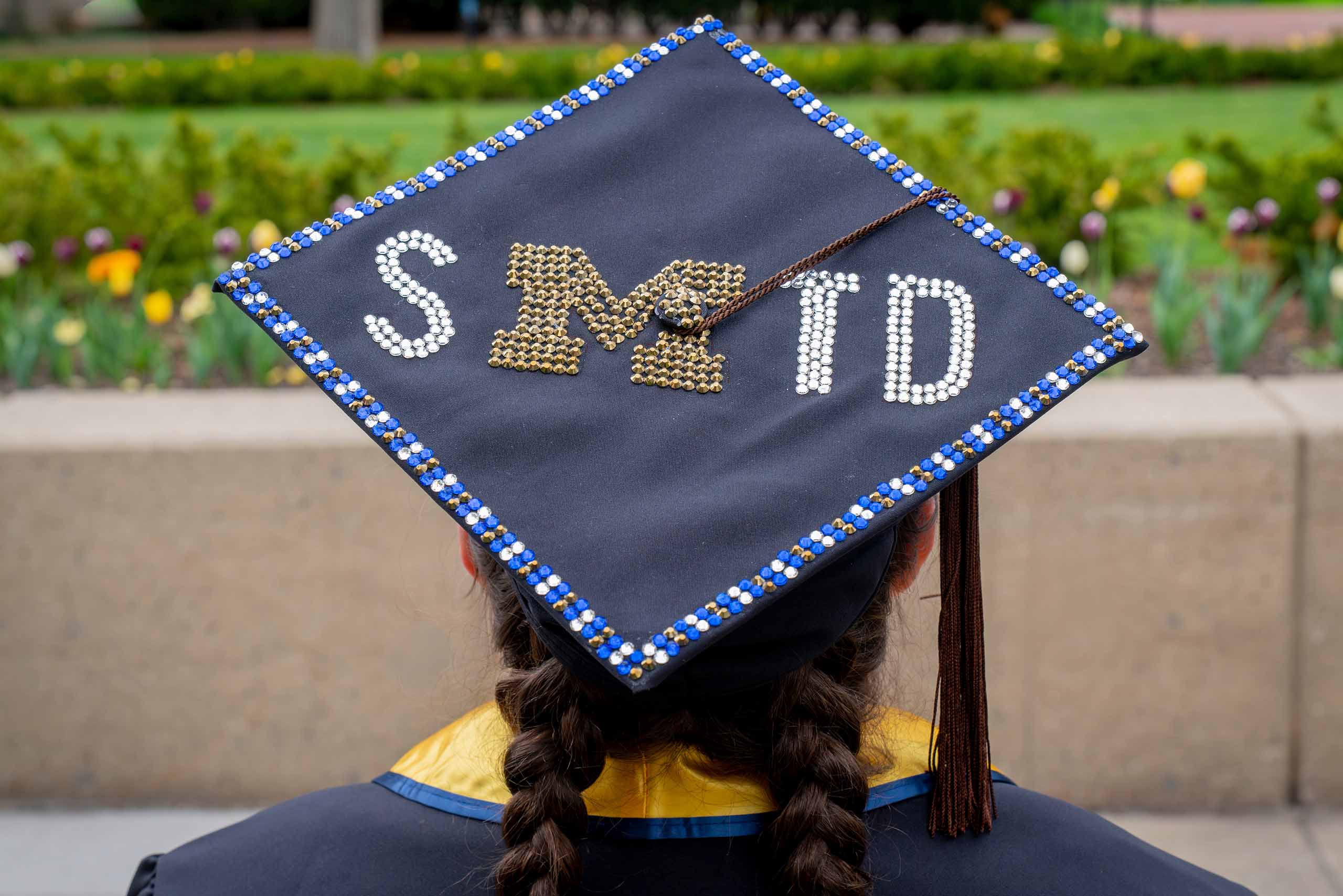A graduate wears a mortarboard cap with "SMTD" decorations added to the top.