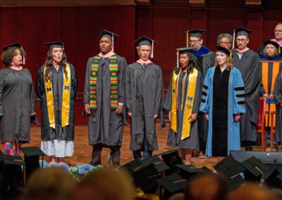 Six graduates perform singing together on stage, with more graduates seated in the audience wearing mortarboard caps.
