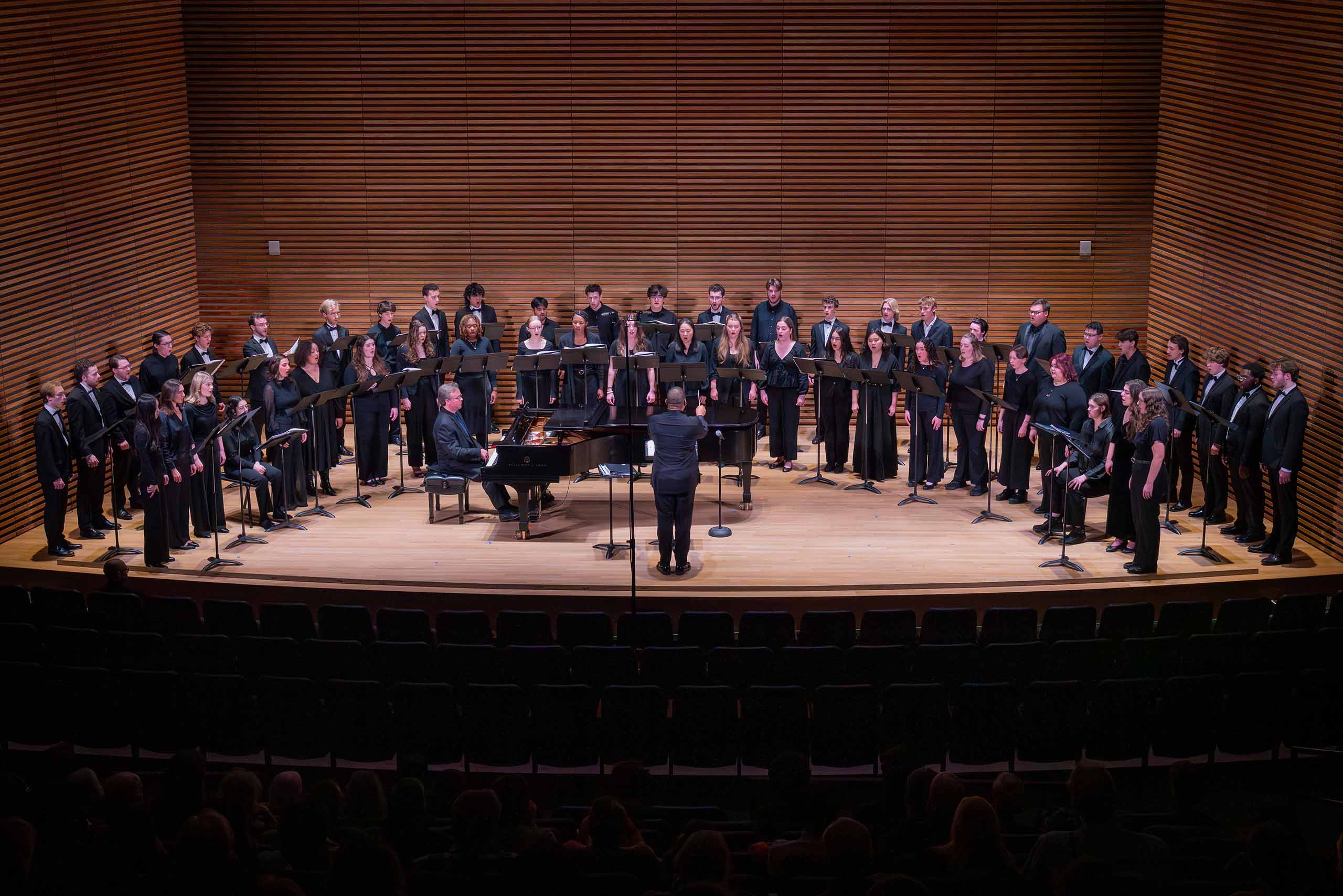 A choir performs on stage with a conductor and pianist, wearing concert black.
