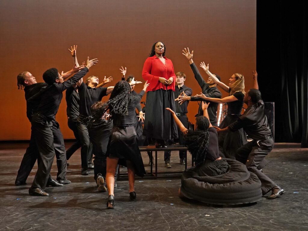 On a studio stage, one performer stands on a riser wearing red, while a group of other performers all wearing black surround her with choreographed, outreached arms.