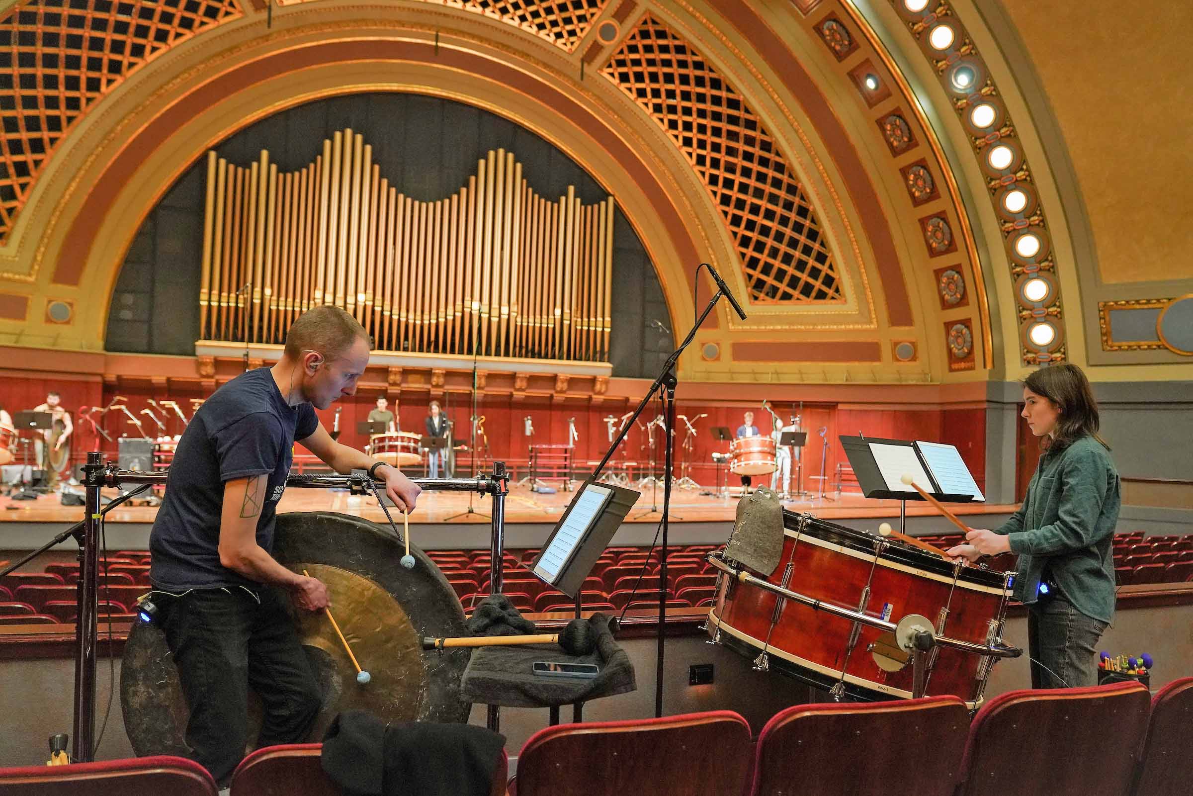 Two large drums are played in the house of Hill Auditorium, with more percussion and recording equipment seen on stage in the background.