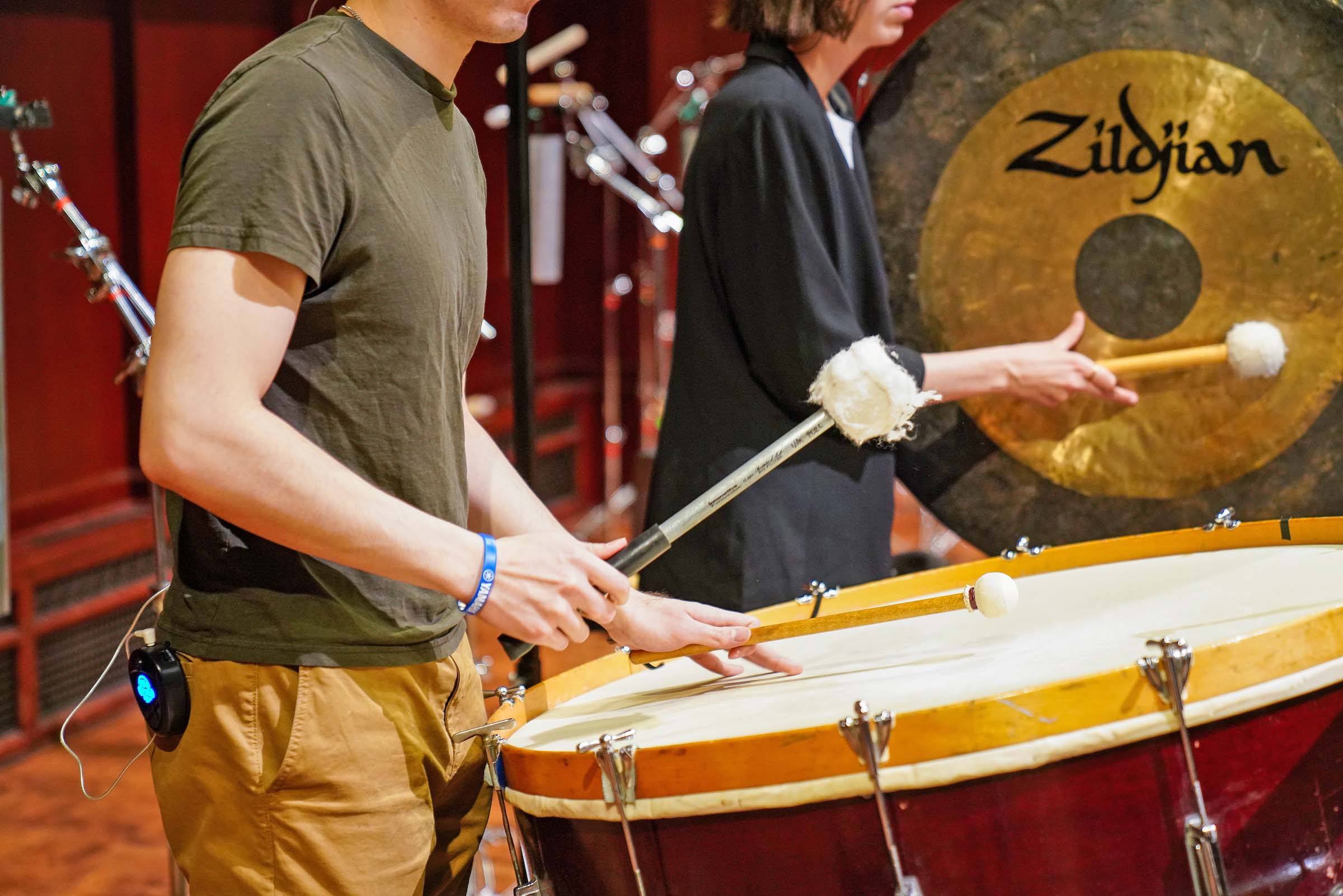 Two percussionists play a timpani drum and a gong side-by-side, standing on a stage.