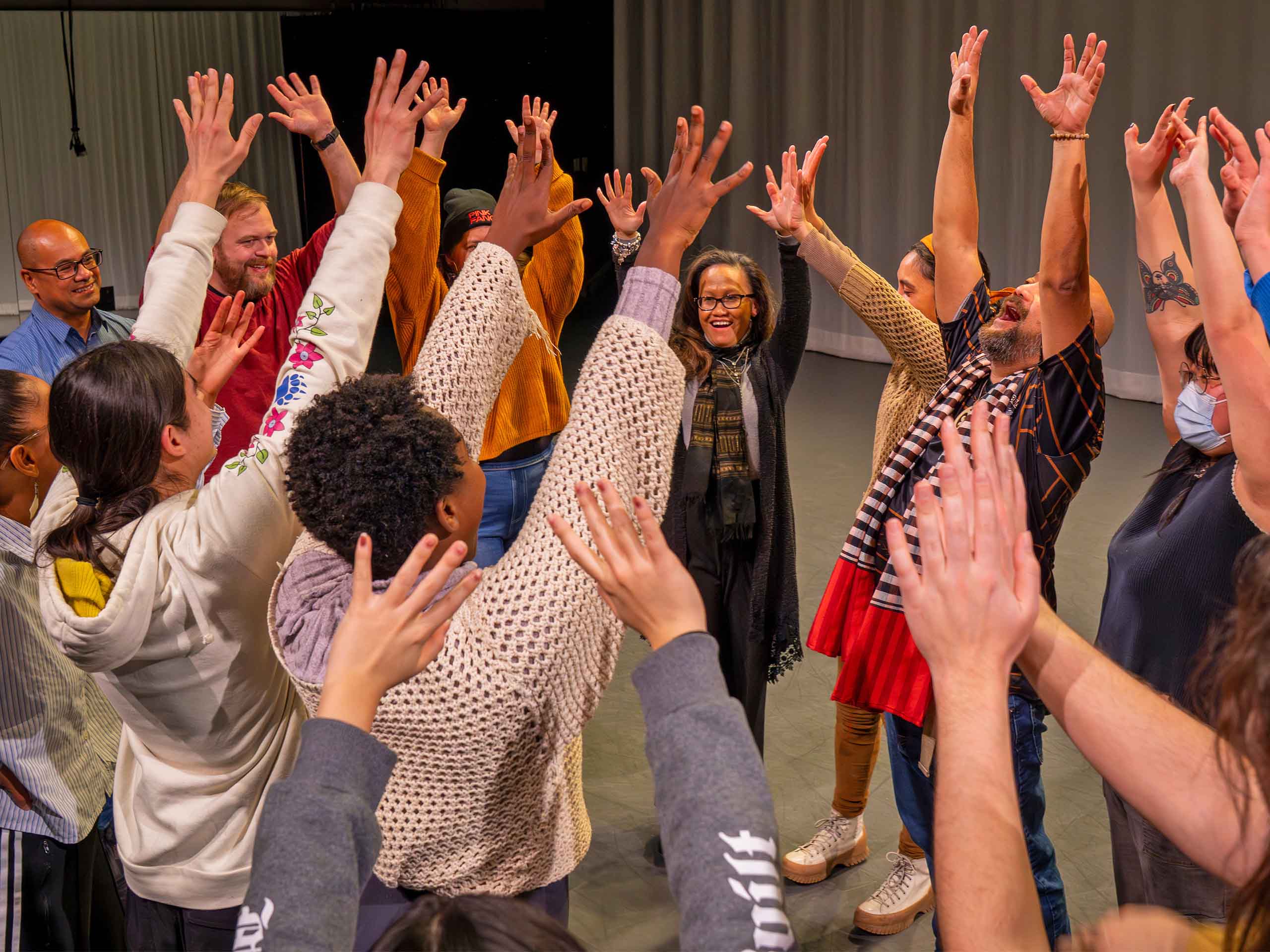 In a studio space, at least a dozen people stand in a close circle with their arms raised.