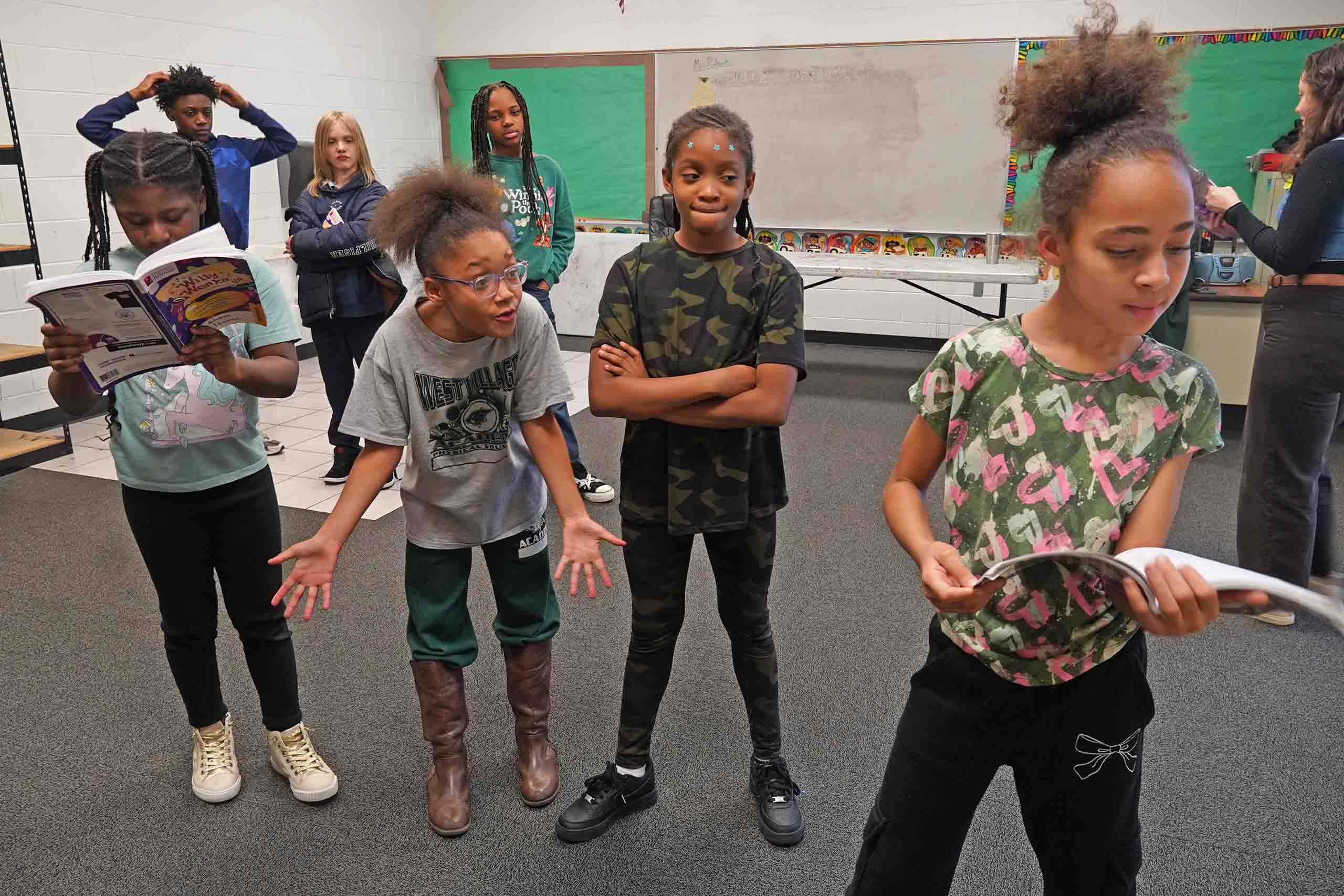 In an elementary classroom, young students stand holding scripts and acting in character.