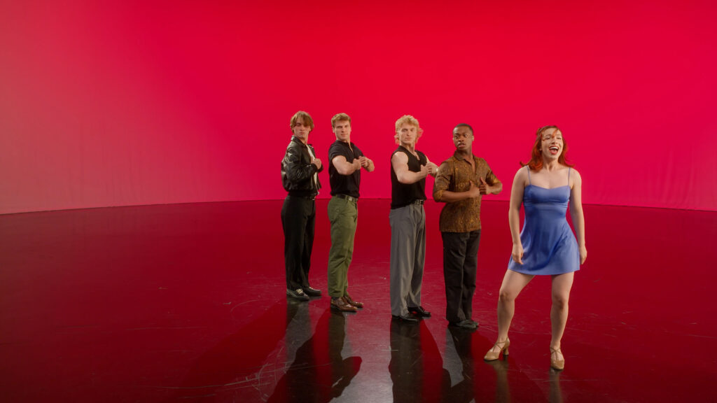 On a stage with vibrant red backdrop, a lead singer performs, with four other actors posed in a straight line behind her.