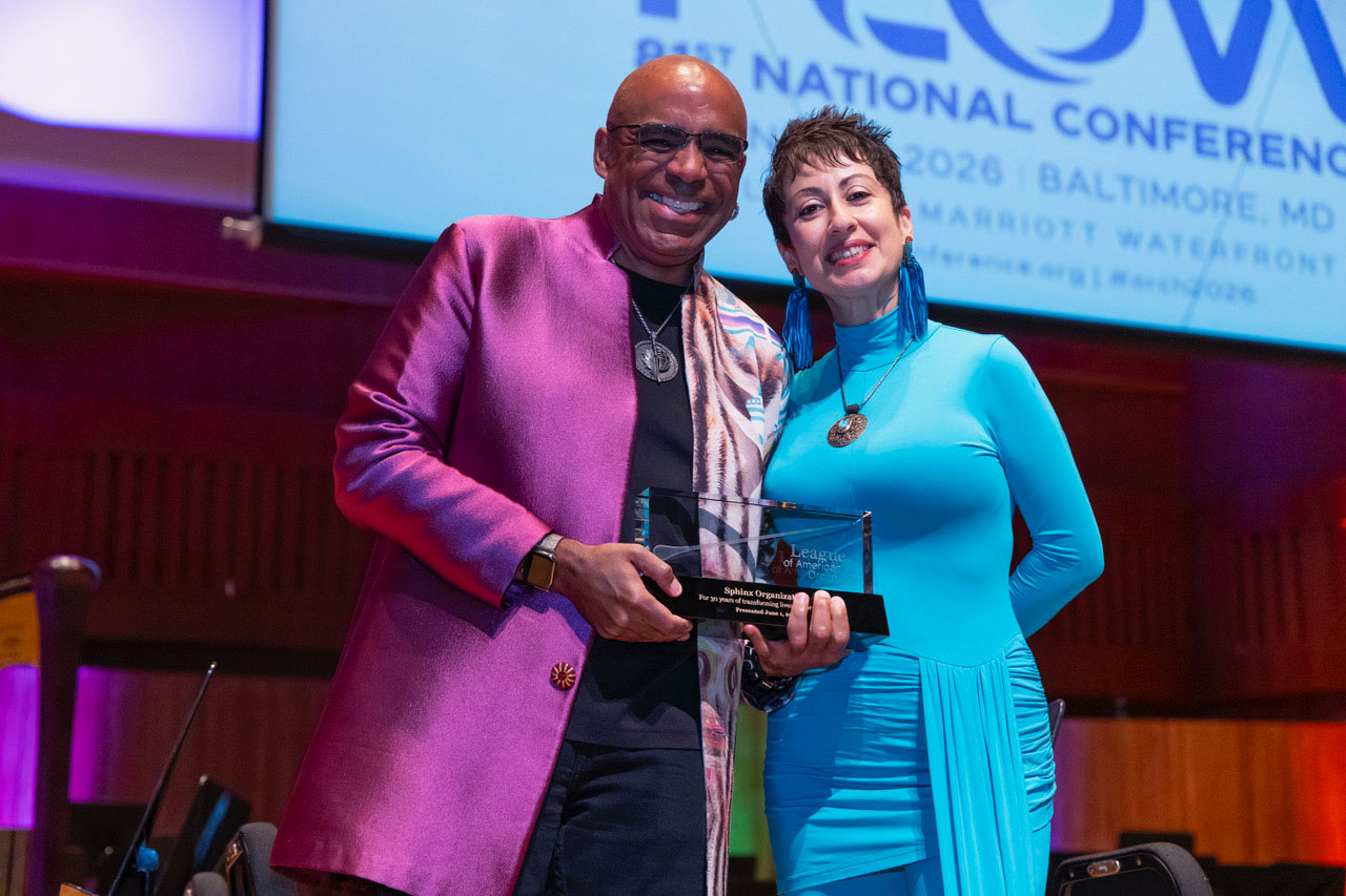 Aaron and Afa Dworkin pose standing together on stage with a glass award. Aaron is attired in a purple suit coat, and Afa wears a bright blue dress and earrings.