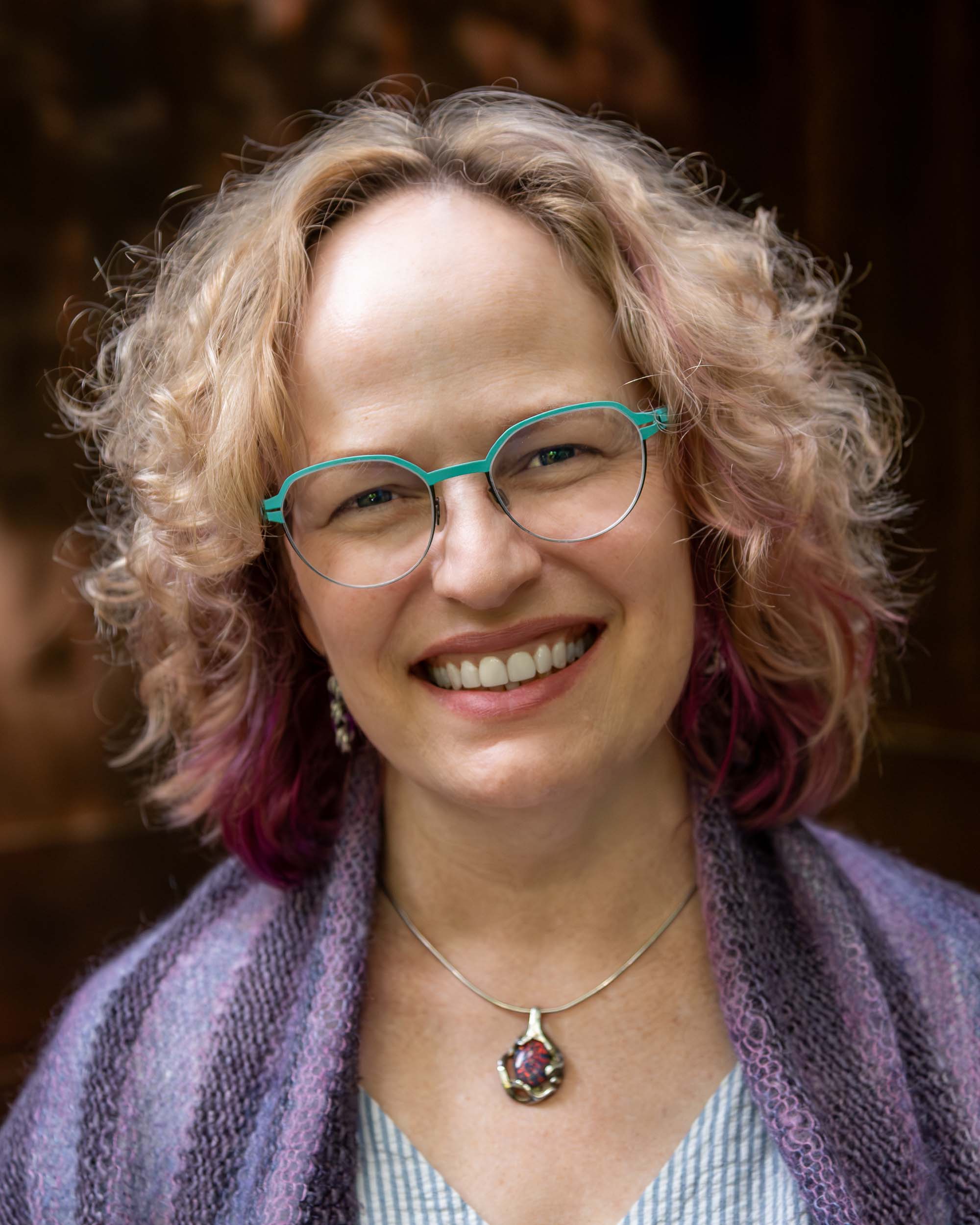 Headshot of Amy E. Hughes, wearing teal-rimmed glasses, a purple shawl, and a necklace.