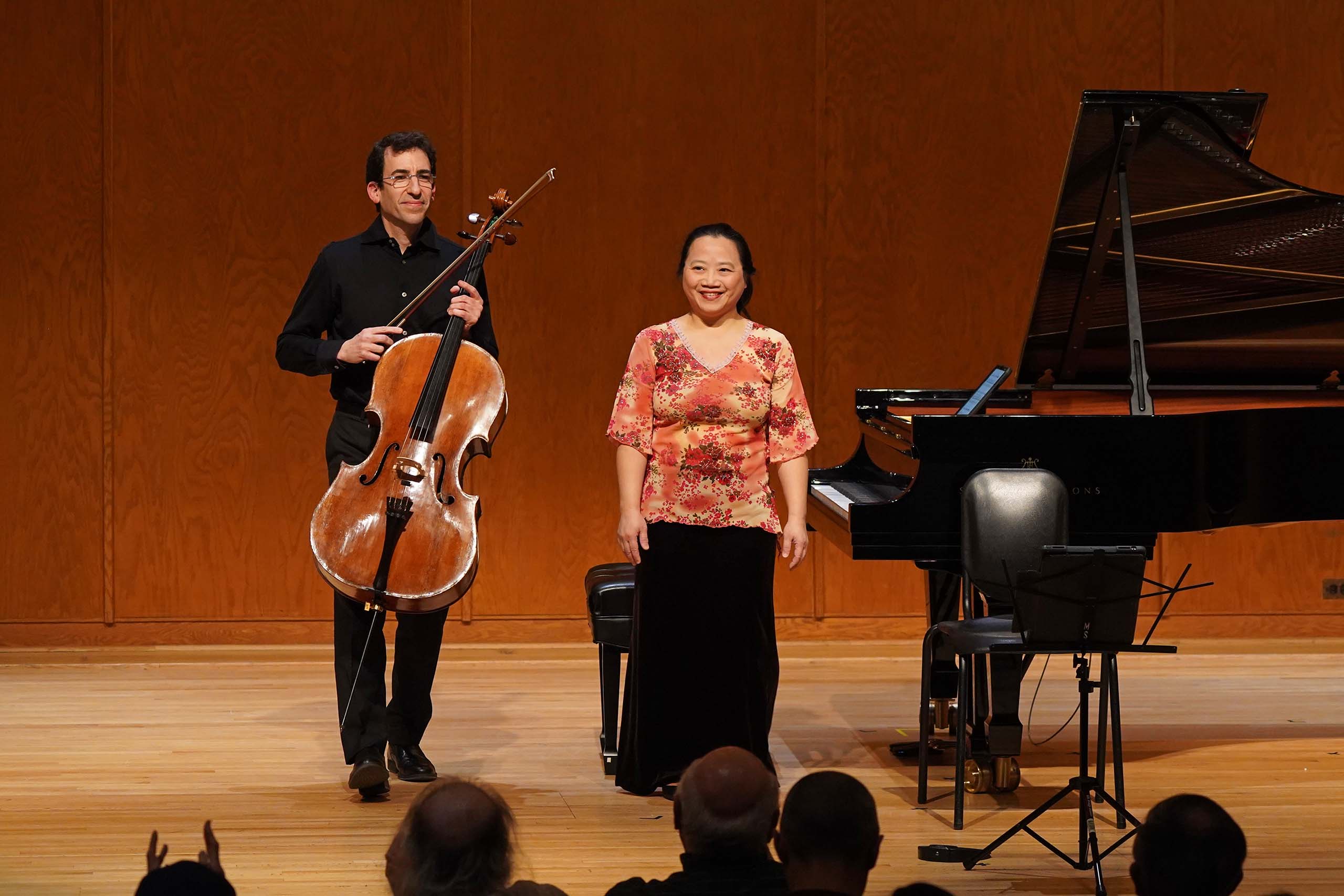 On a recital hall stage, Cheng stands next to a grand piano and Eldan stands holding his cello, with audience heads visible in the lower foreground.