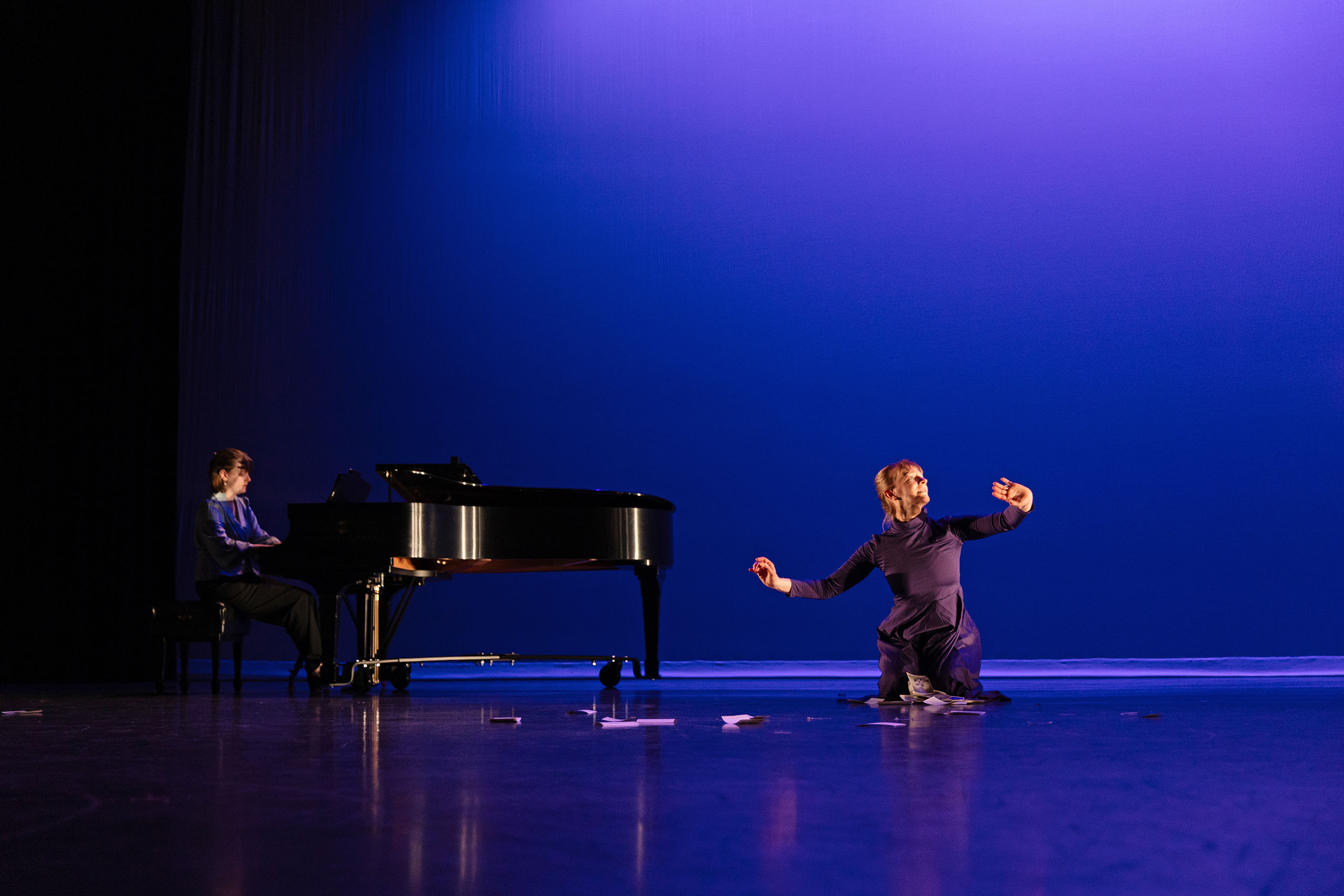 On a stage with purple backlighting, a dancer costumed in black performs with wrists held out and scattered papers. Nearby a pianist plays on a grand piano.