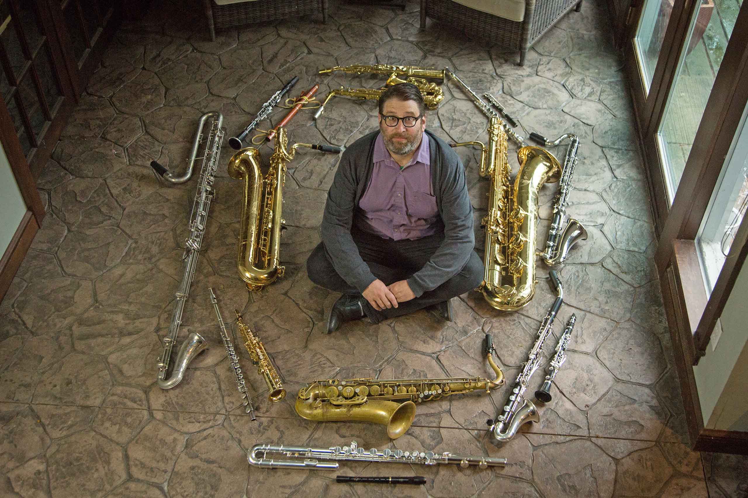 Andrew Bishop is seated cross-legged on a stone tile floor, looking up at the camera a floor above. He is surrounded by about 16 saxophones and other wind instruments of various sizes laying on the floor.