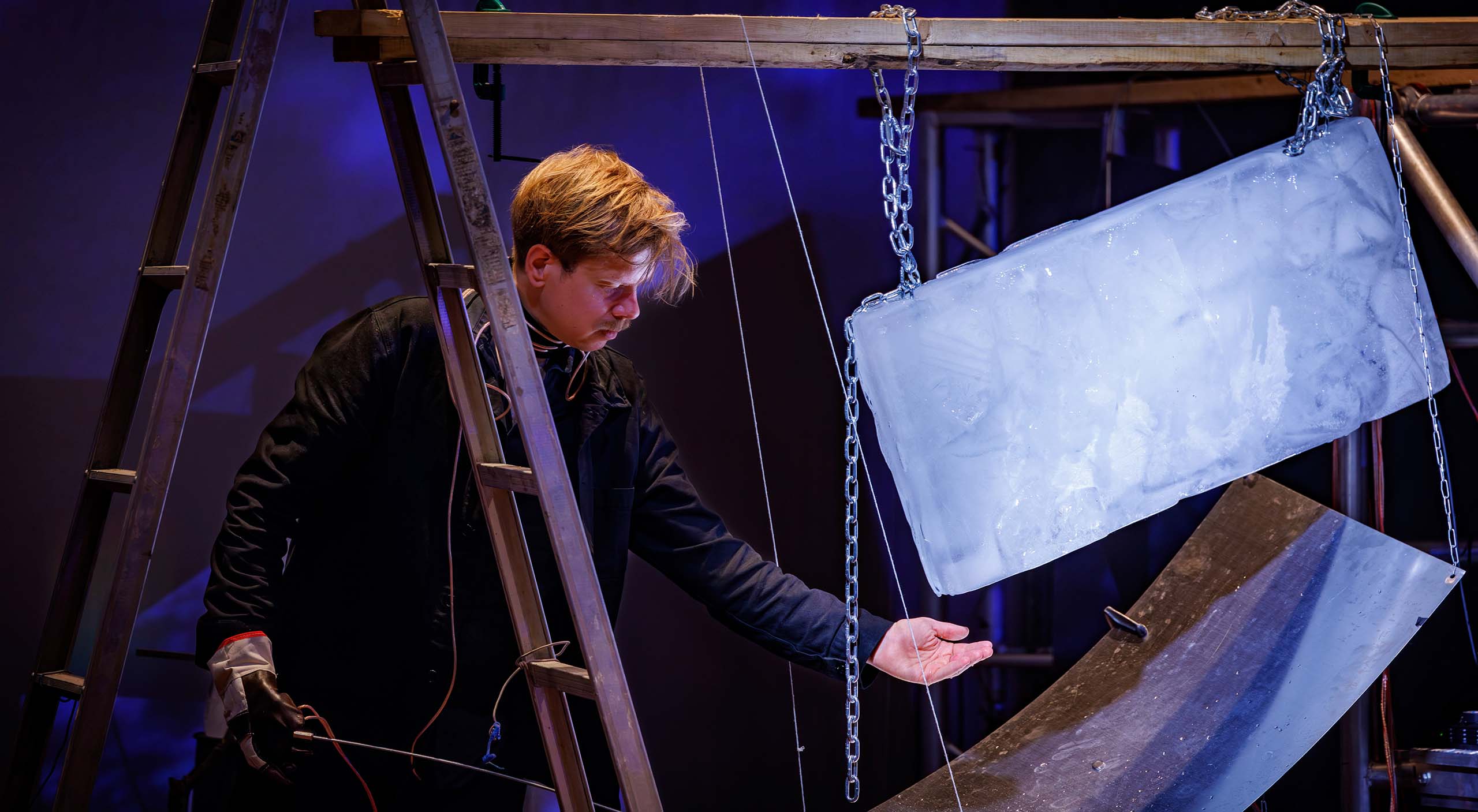A large block of ice hangs from chains, held up by boards set across a ladder and scaffolding. A man wearing black holds his palm out to catch drips off the corner of the ice block.