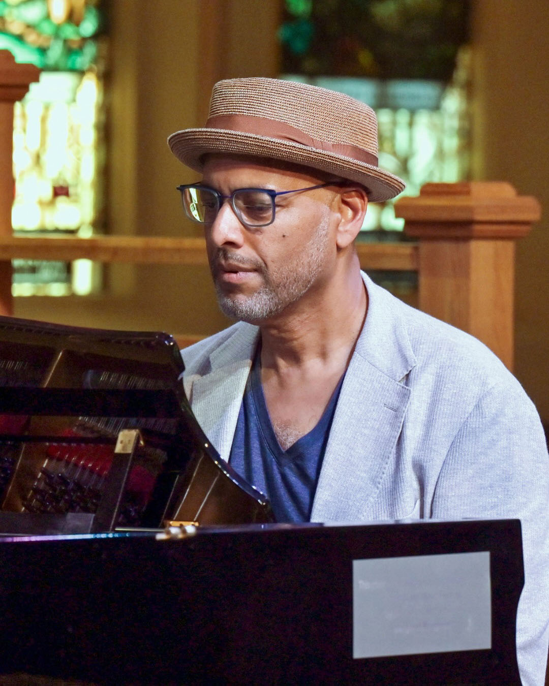 Andy Milne performs seated at a piano, attired in a light blue jacket and weaved brown boater hat, with stain glass windows in the background.
