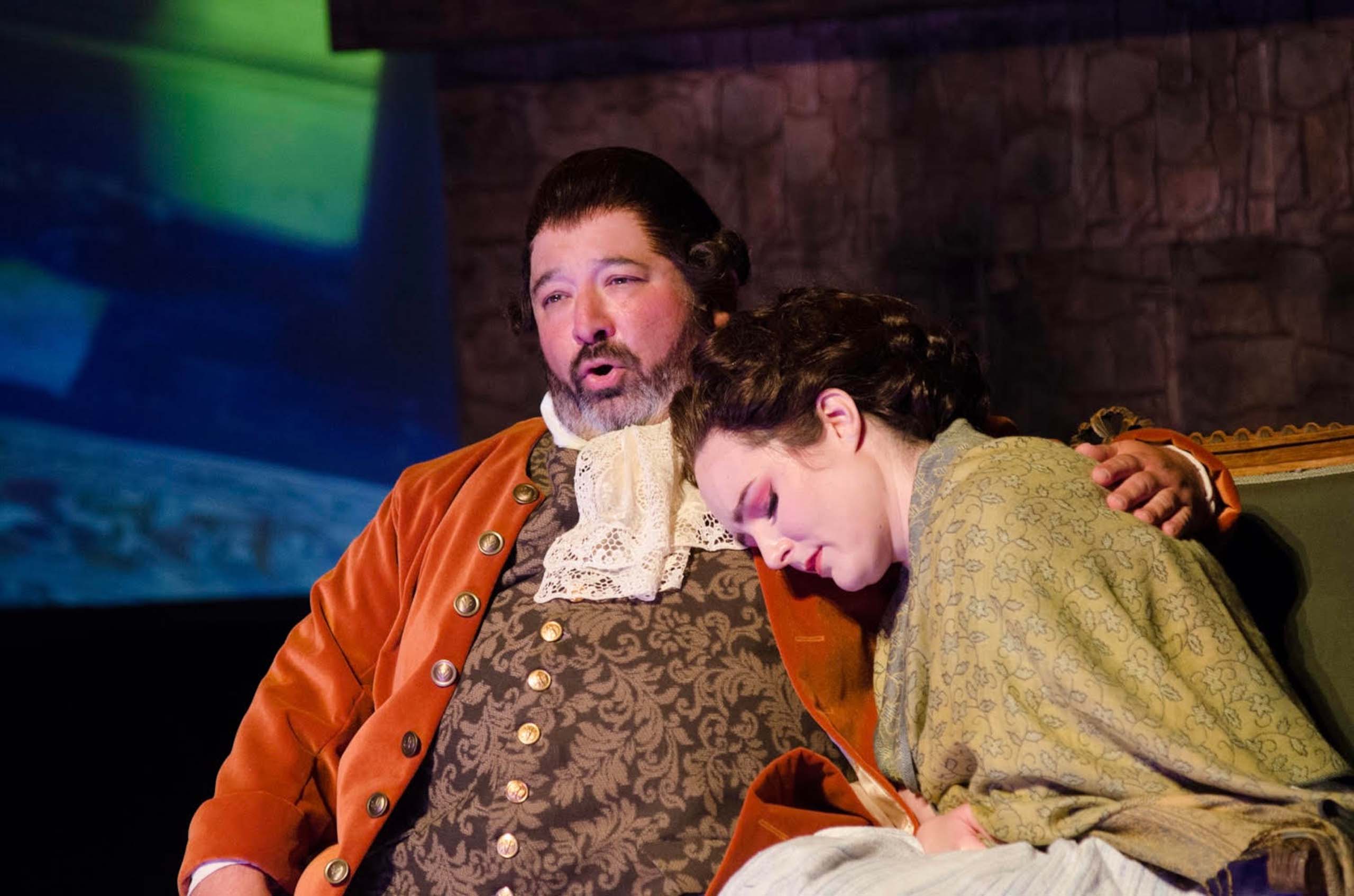 Two actors perform a scene on stage seated in a relaxed embrace, as Andy Papas' character sings and the other has her eyes closed. They are costumed in 18th century attire.