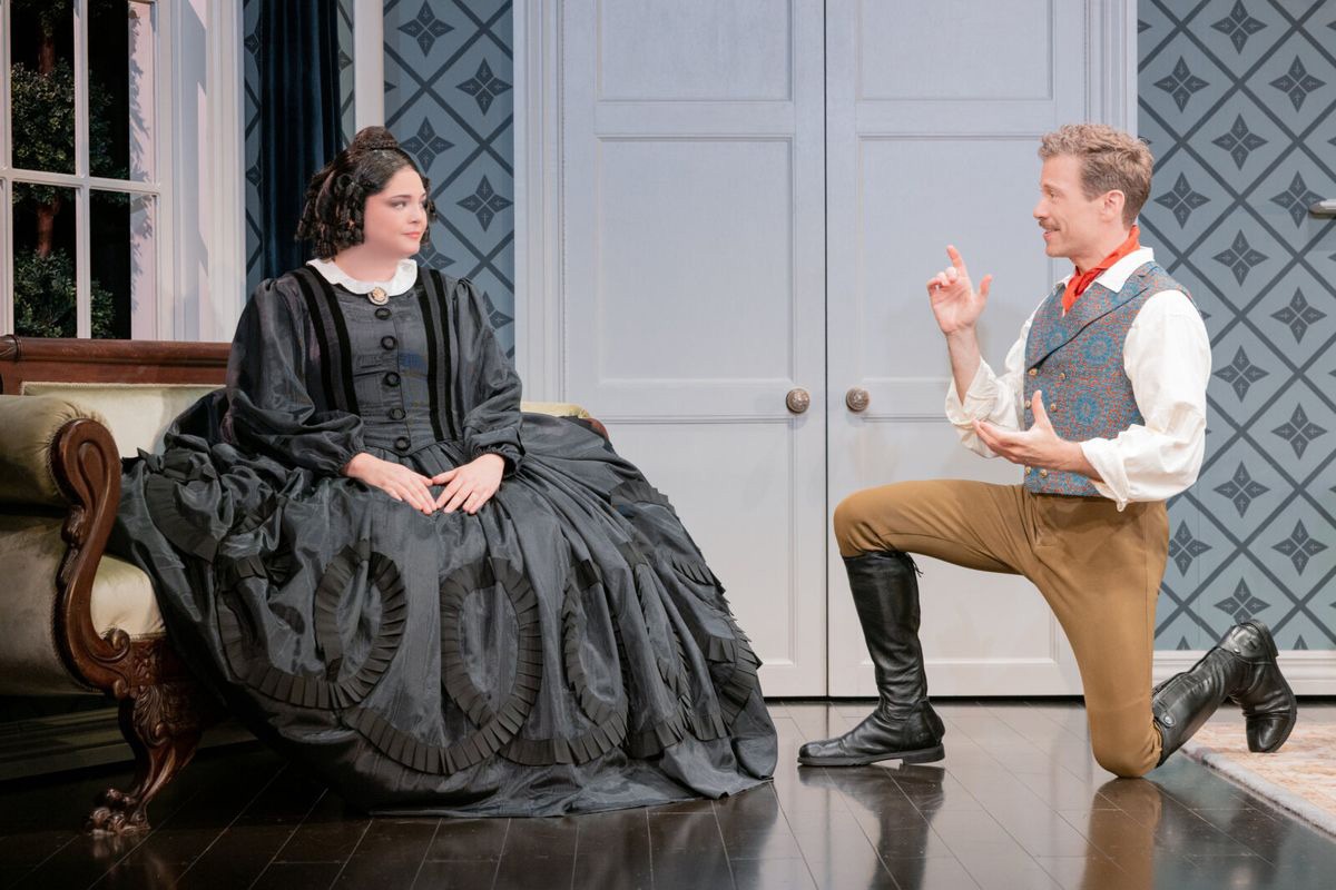 Two actors perform on stage, costumed in 19th century attire. One is seated, wearing a long black dress, while the other kneels on the ground before her.