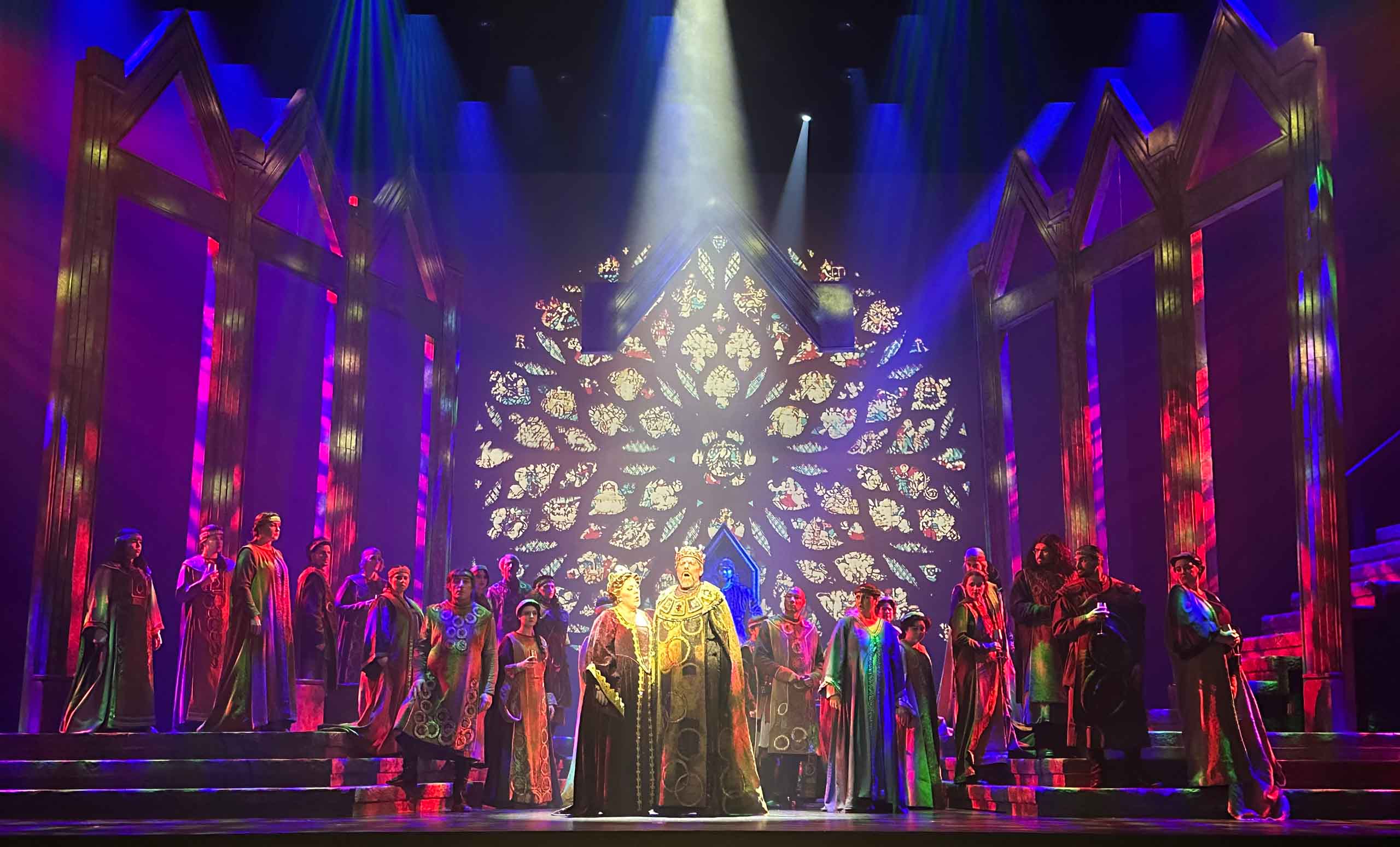 A cast of about 2 dozen performs on stage, with a singing royal couple in long robes centered under a spotlight. The set is a historic cathedral scene, with a huge intricate, round stain glass window. The set is dramatically lit with red and blue lighting.