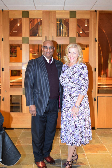 Two people pose standing together in a church lobby, smiling, wearing formal attire.