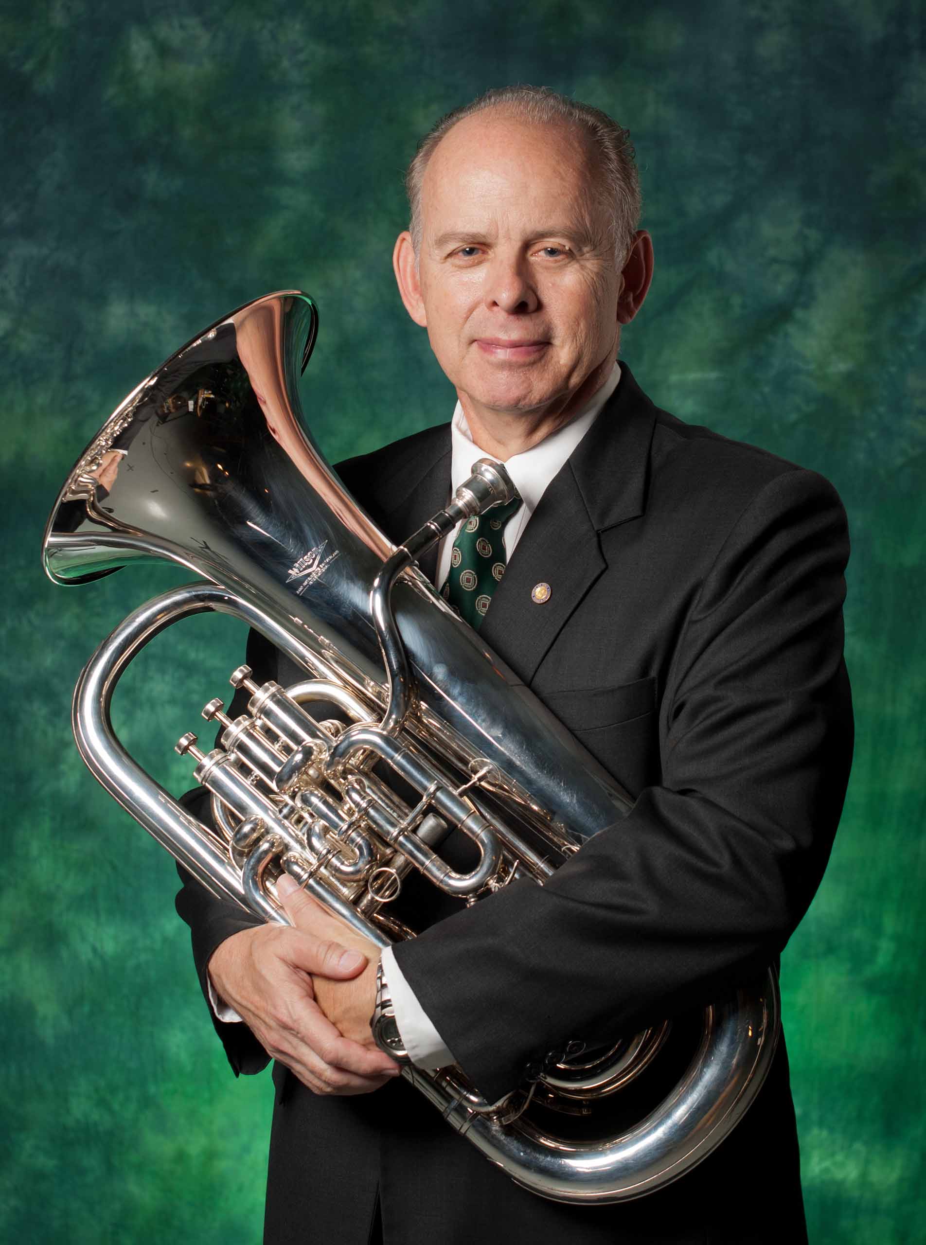 Formal standing portrait of Brian Bowman holding a silver euphoium in a suit, with a green backdrop.