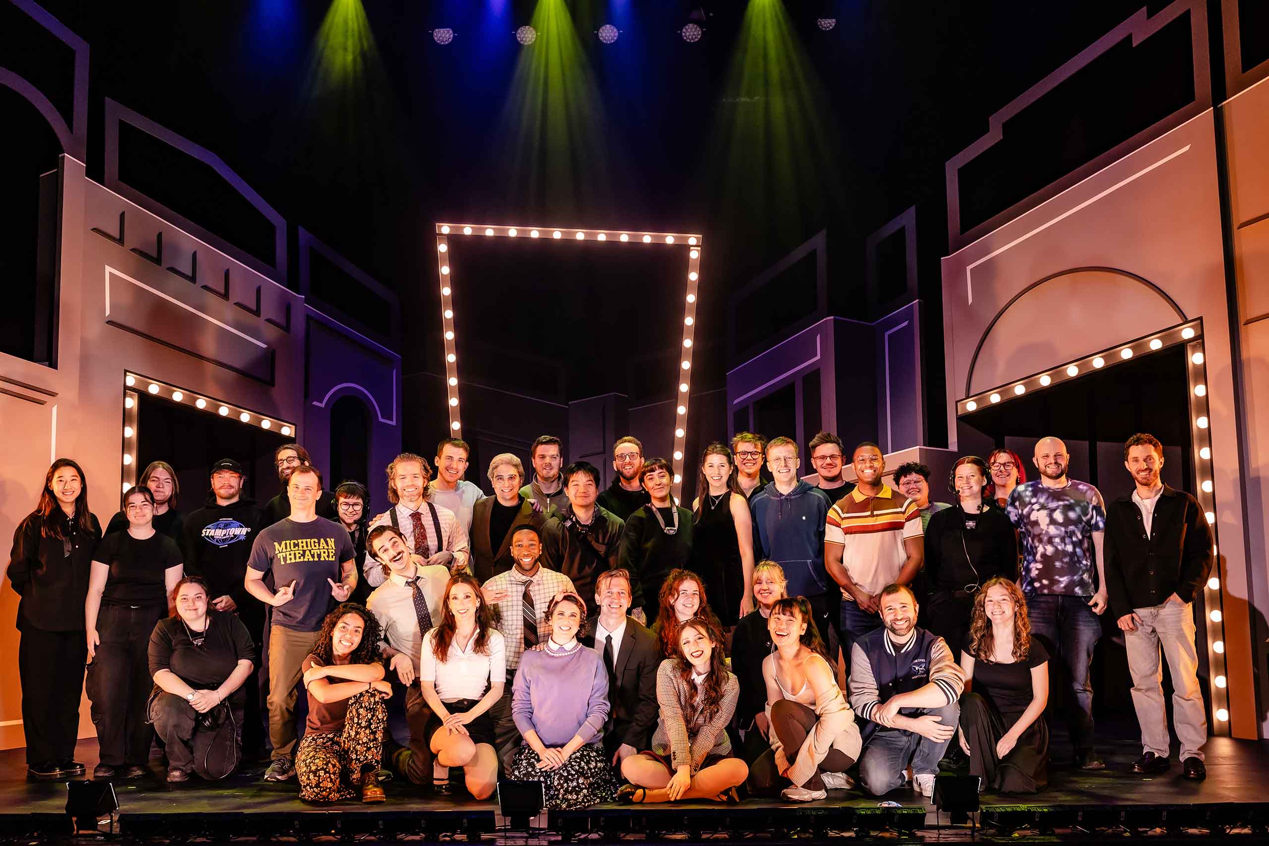 About 3 dozen cast and crew members pose together in several rows on stage. Behind them, the set features sleek white buildings and doorways framed with marquee bulbs. Brian Holden points with both hands to his Michigan Theatre t-shirt.