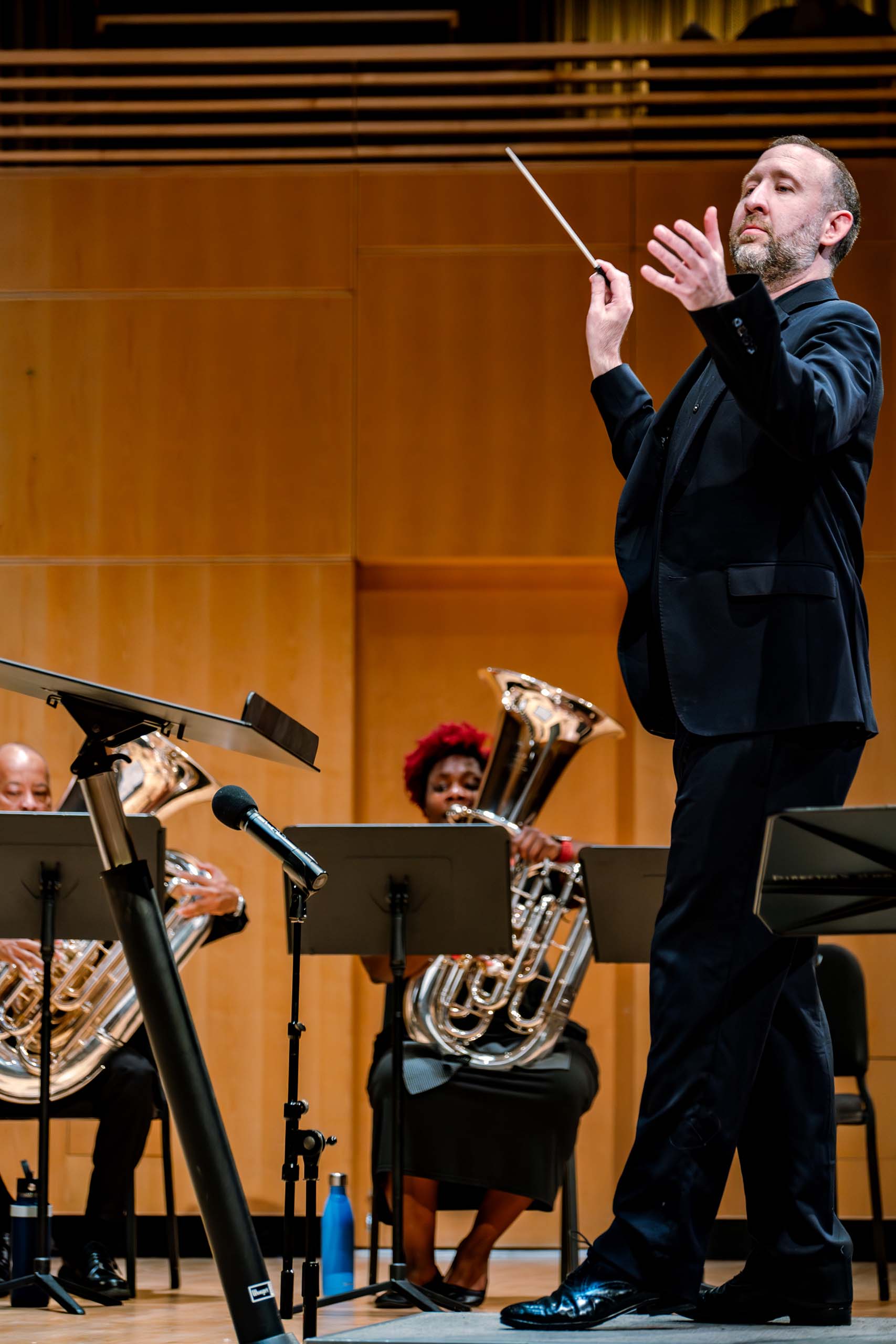 Brian Kaufman conducts standing on a podium, with baton tipped up, wearing dark velvet concert attire. Two brass players in his ensemble are seen seated in the background.