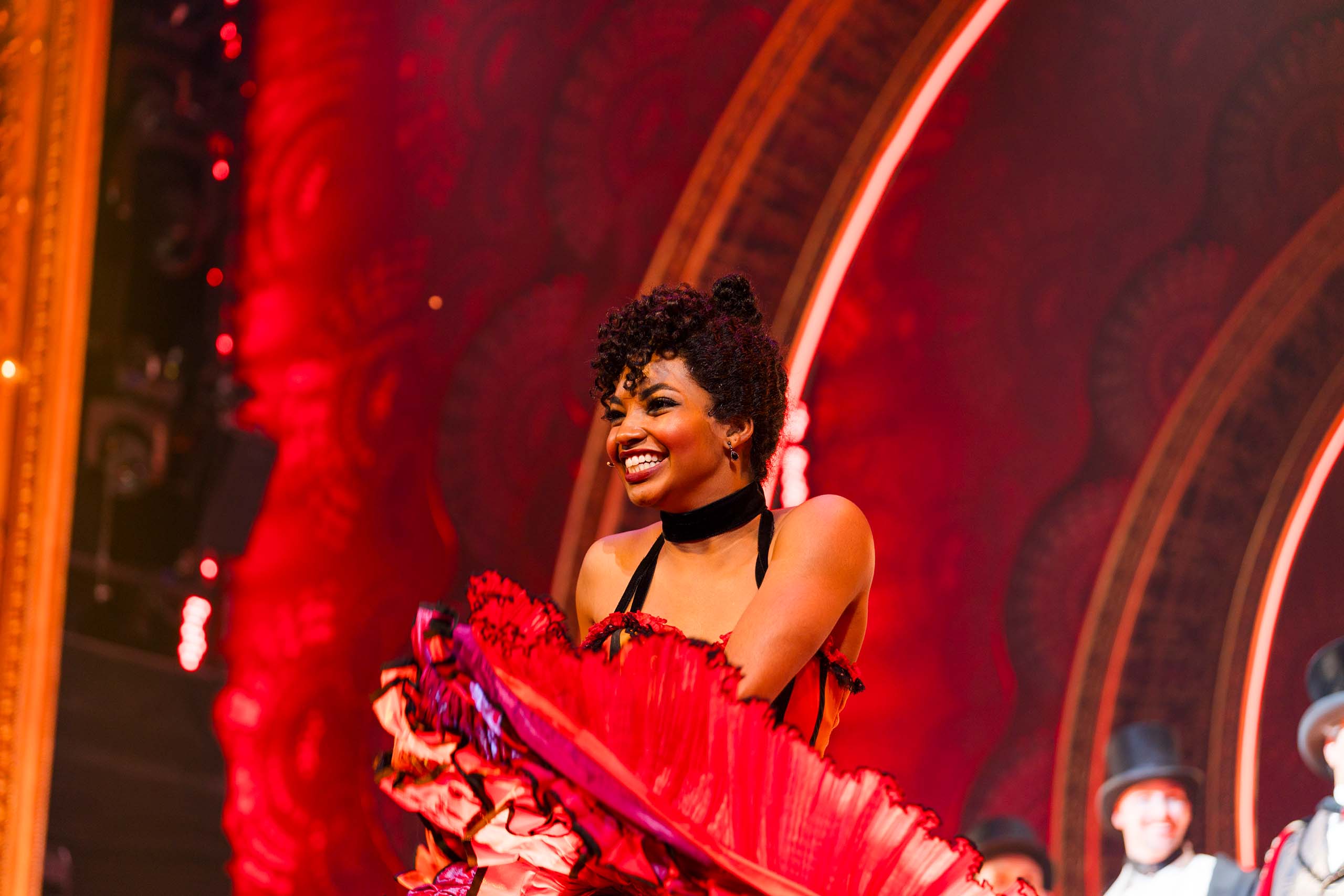 Brooke Taylor dances in a performance on stage, costumed in a frilly red dress with black straps and neckband. Castmates in top hats and dramatic theatre arches from the set are in the background.