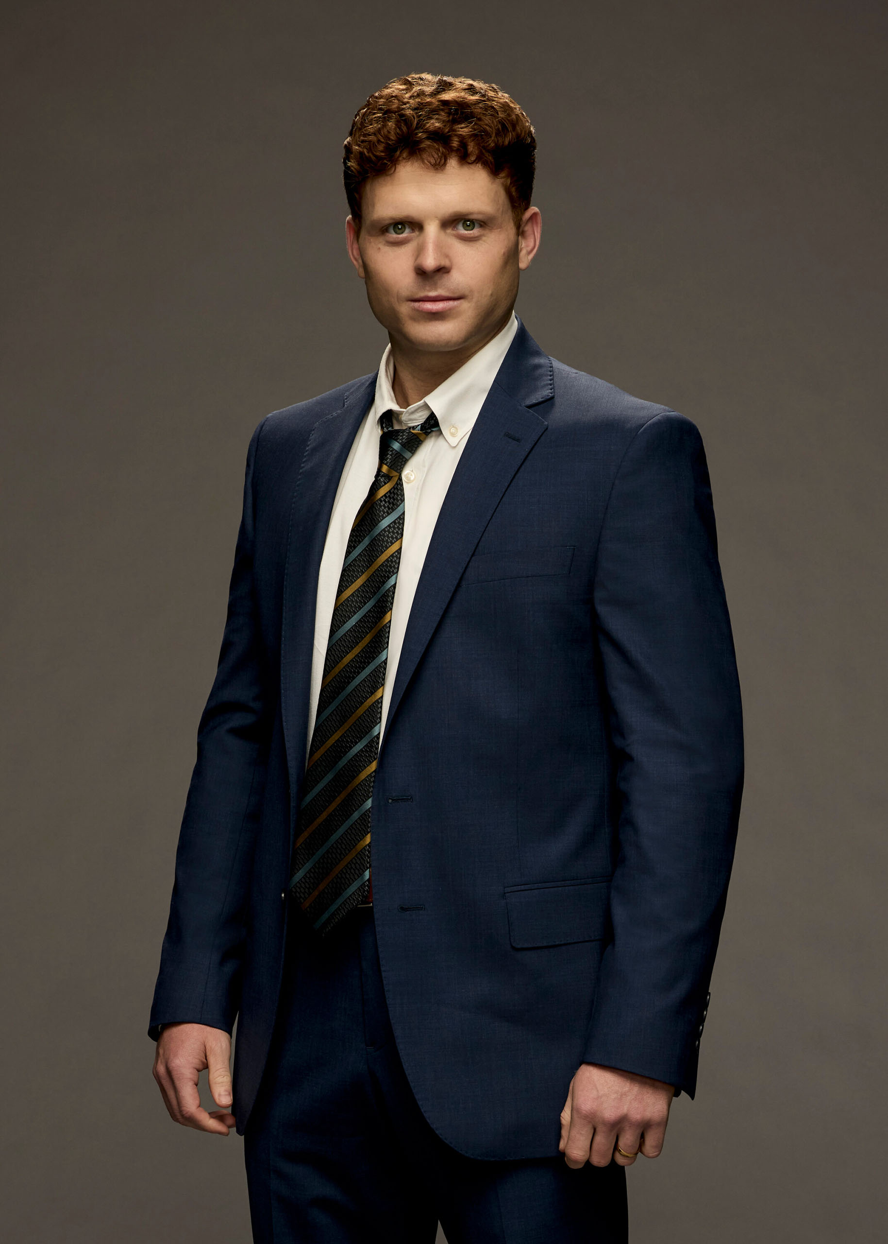 Studio portrait of Caleb Foote in character, standing costumed in a navy blue suit and striped tie.
