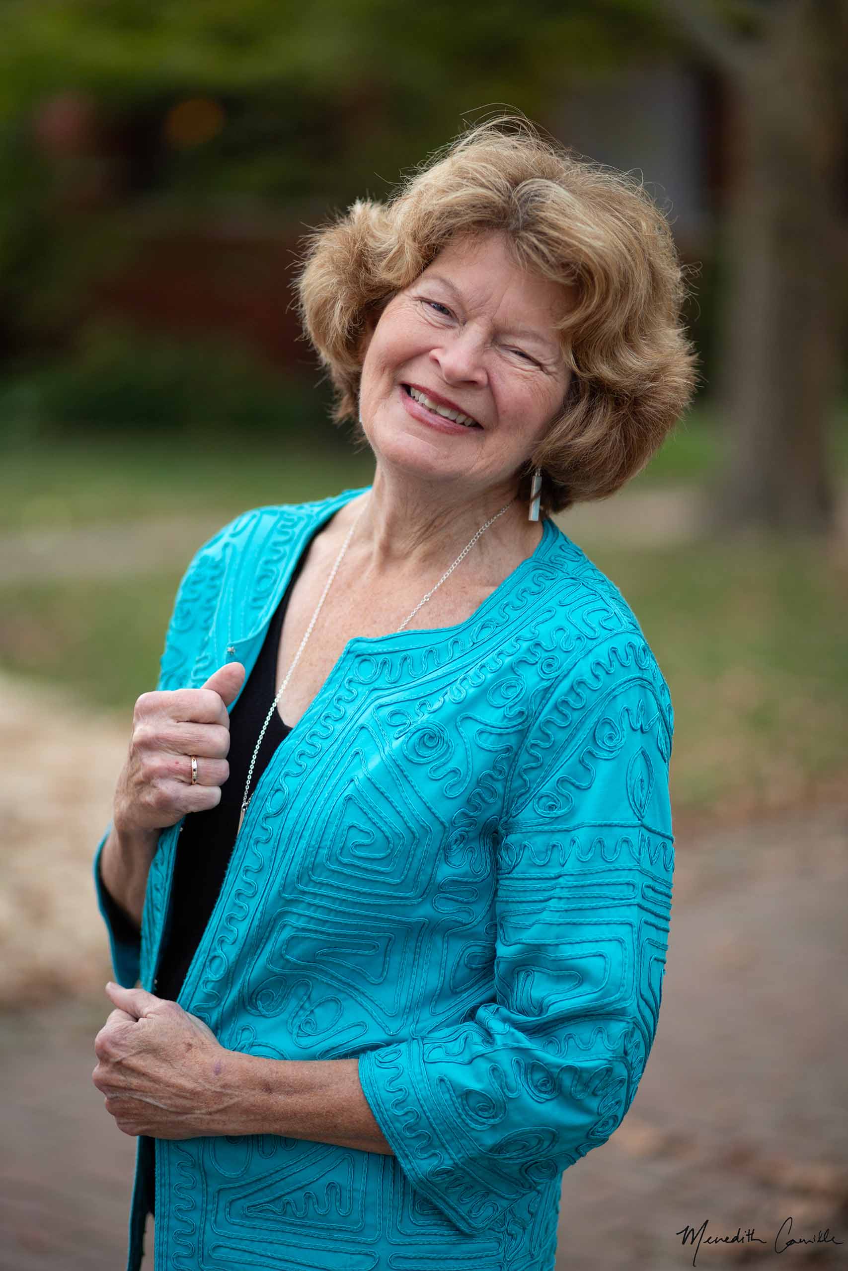 Standing portrait of Catherine McMichael smiling, with hands gripping the edge of her patterned teal jacket.