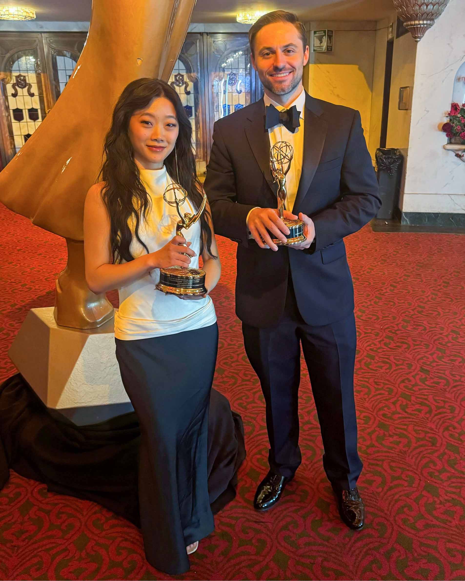 Catherine Miller and  Patterson McKinney pose standing together in an elegant space in formal dress, each holding an Emmy Award.