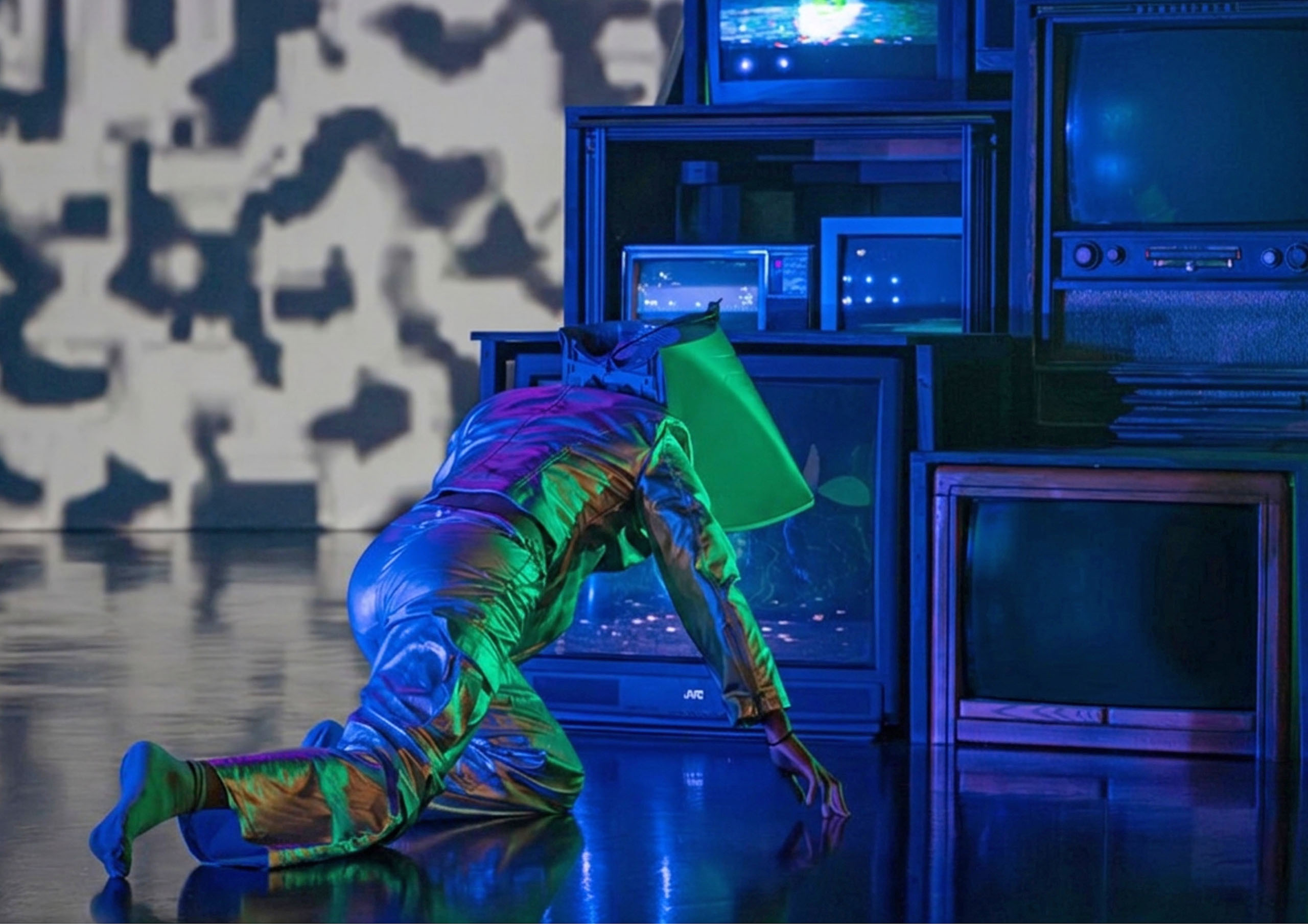 A dancer crawls near a grouping of televisions, in futuristic silvery costume.