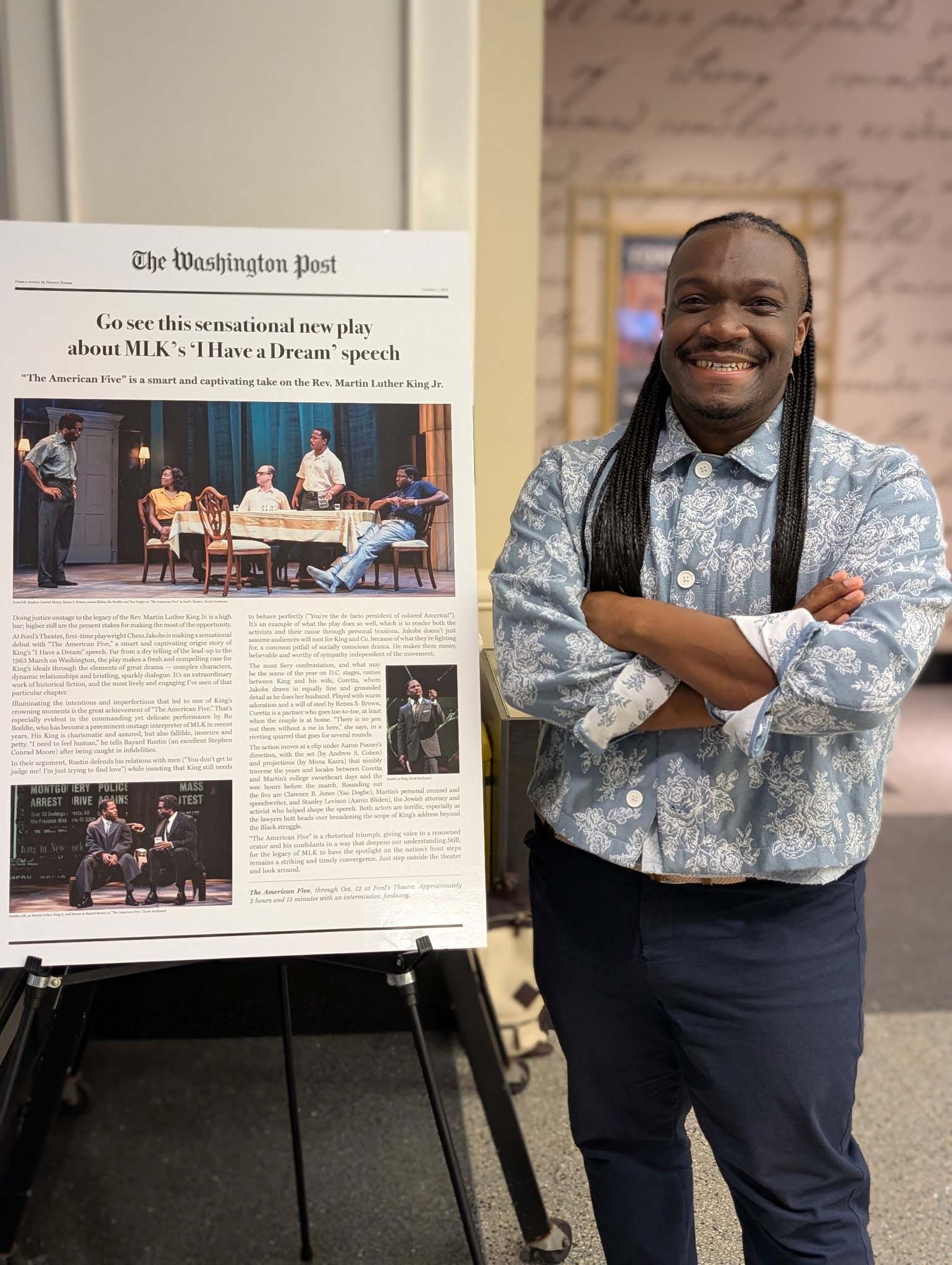 Chess Jakob poses standing next to a large mounted newspaper article on a stand, with arms folded. The headline says: Go see this sensational new play about MLK's 'I have a Dream' speech.