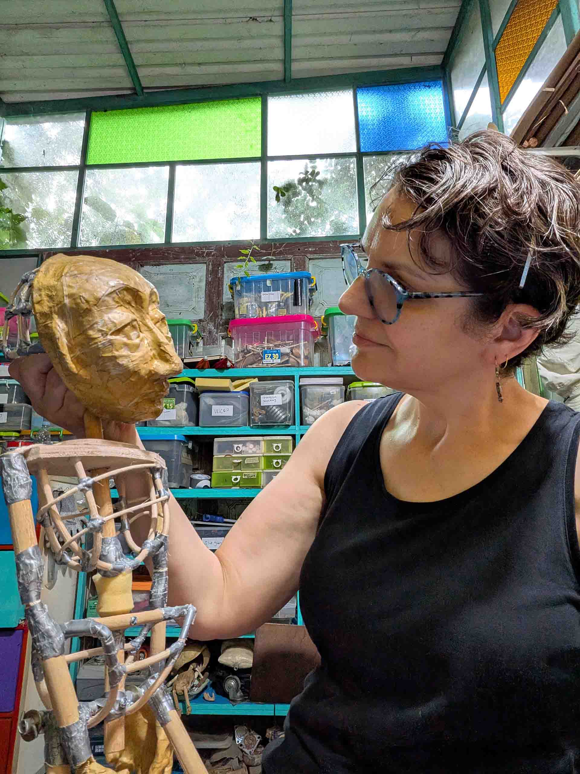 Christianne Myers stands looking at a puppet with detailed face and body framing. Behind her are workshop shelves full of supplies in plastic boxes, and industrial windows above with clear and colored glass panes.