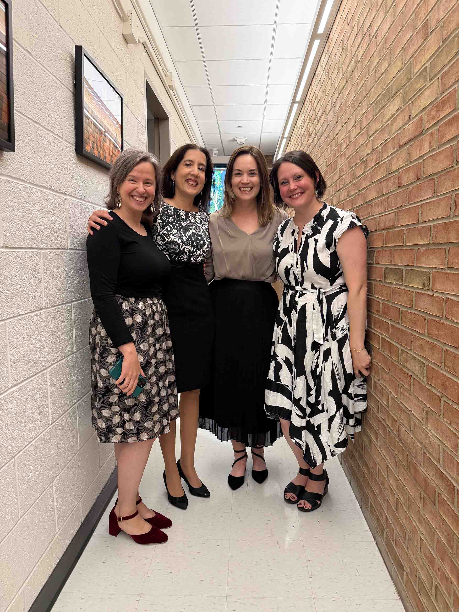 Four people pose standing together in a narrow hallway at SMTD, with walls of cinder block and red brick. They are all attired in formal dresses and skirts.
