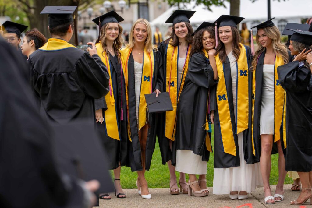 About six graduating students pose standing together outdoors, while someone takes their photo. All are attired in academic regalia.
