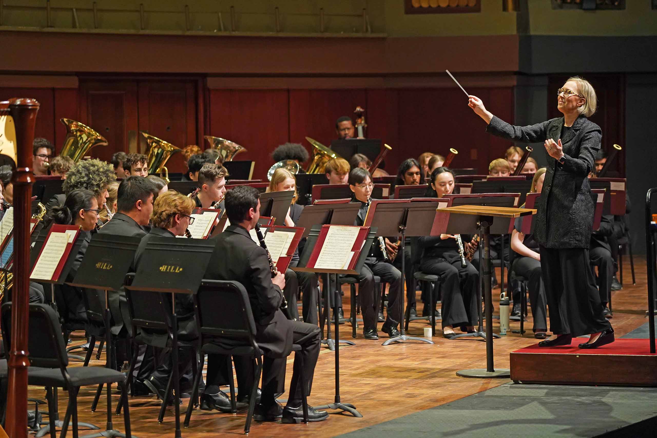 Courtney Snyder stands on a podium conducting a band playing on stage, wearing black formal concert attire.