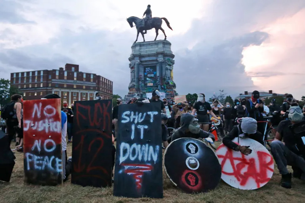 A crowd protests around an equestrian statue on a tall pedestal, many holding signs and wearing masks.