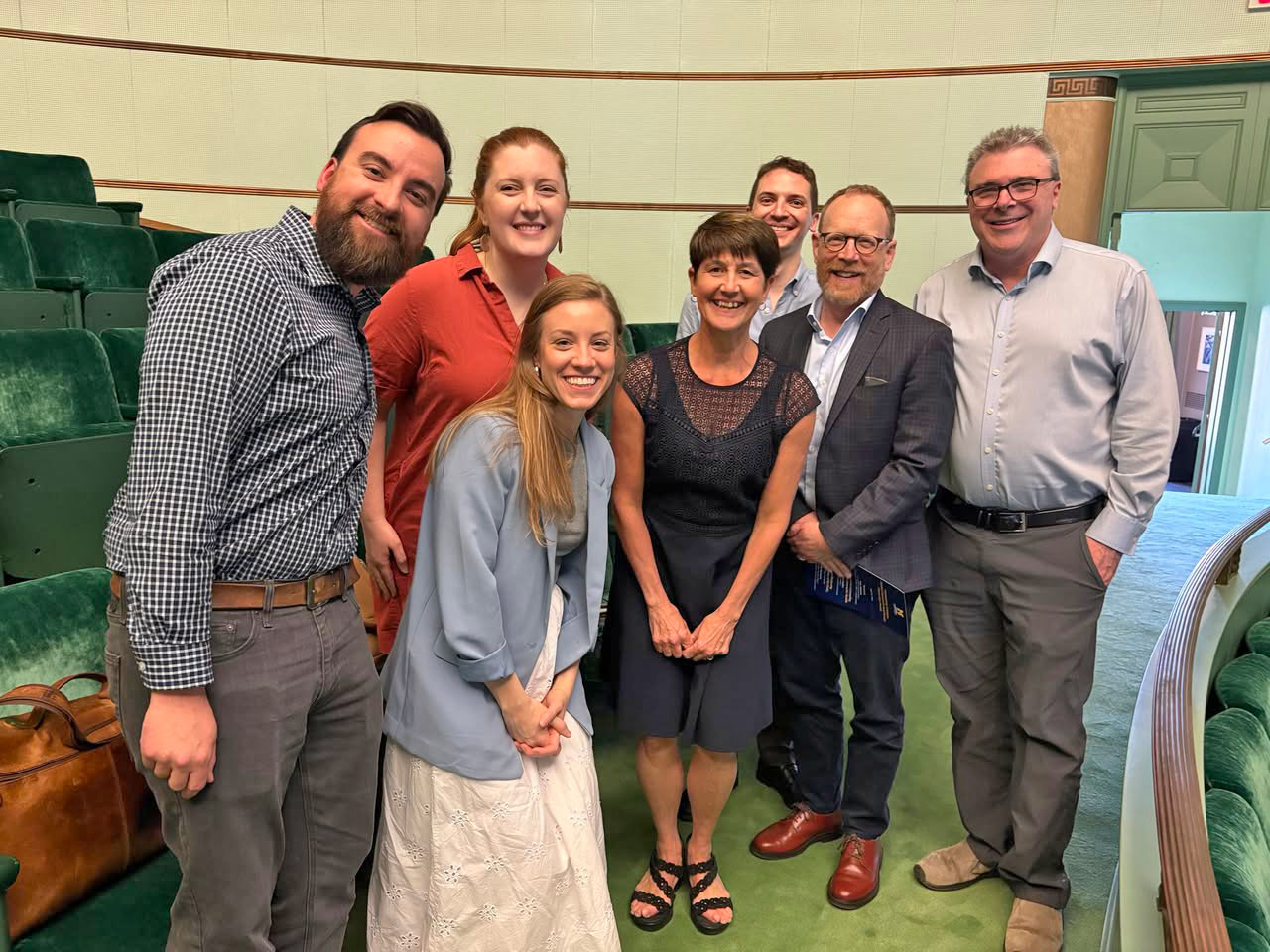Seven people pose standing together in a venue hall, with Colleen Conway at center. All are in business casual attire.