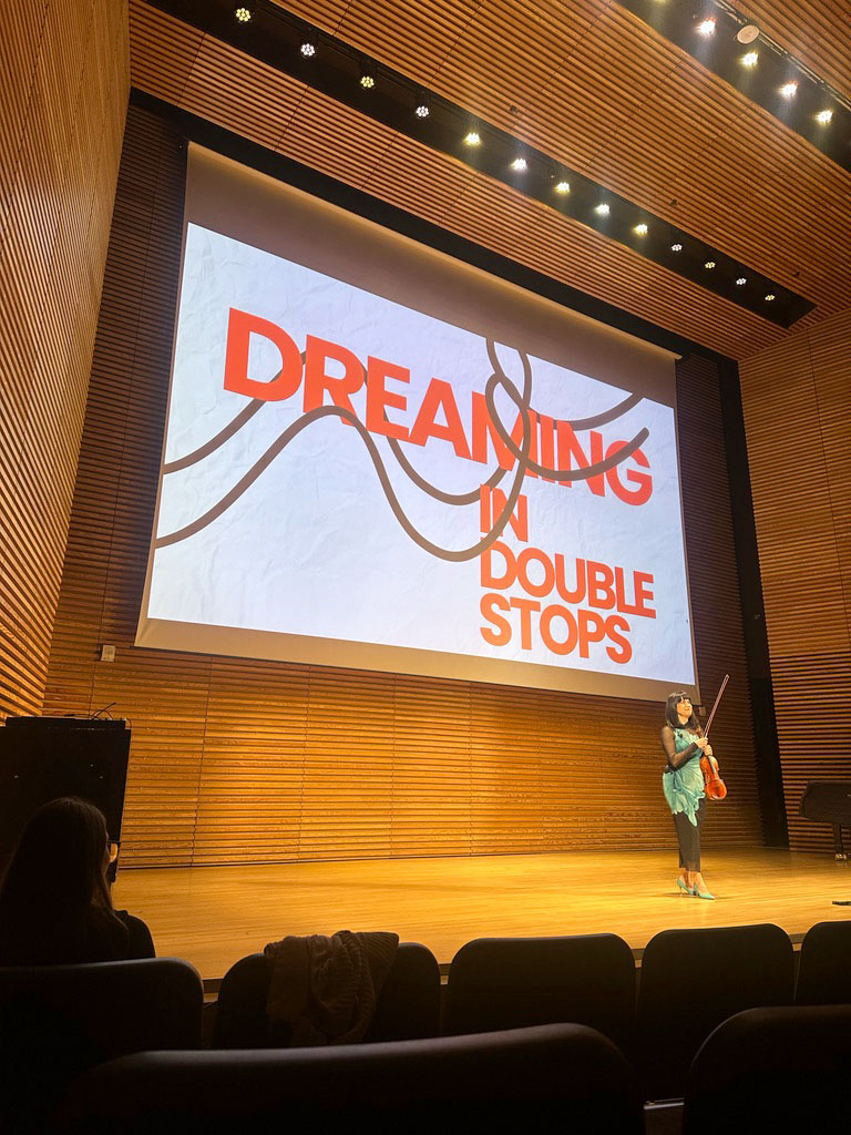 Danielle Belen holds her violin, standing alone on the stage of Stamps Auditorium. A large projection behind her says "Dreaming in Double Stops" in thick red text.