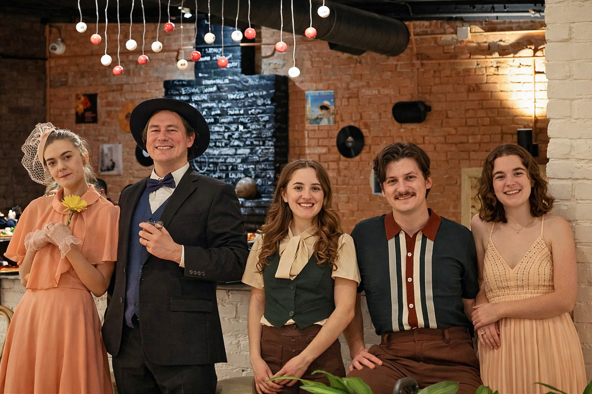 Five people pose standing together in a space with artwork and brick walls. Two are costumed in early 20th century formal attire, while the other 3 are in casual modern clothing.