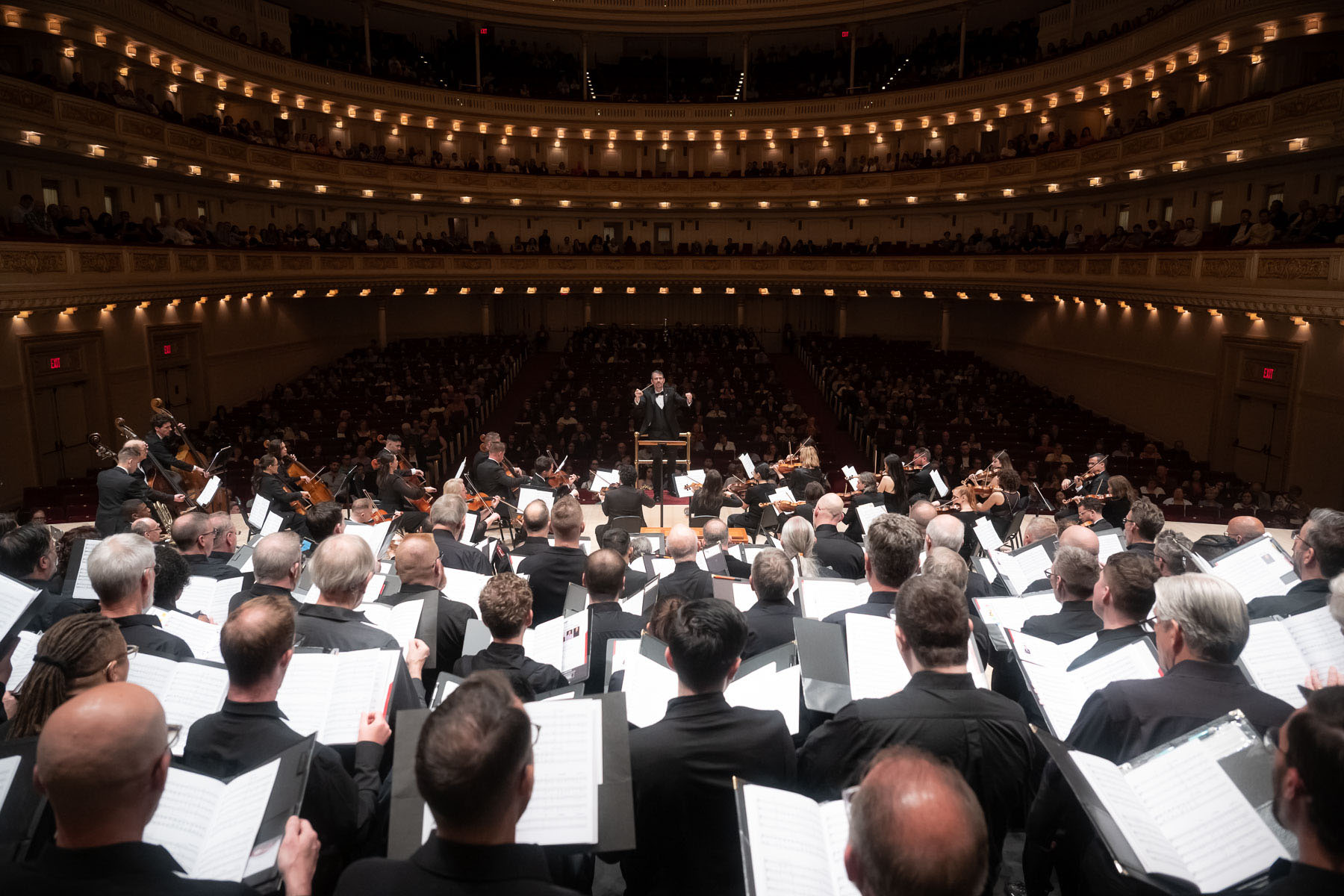 Wide angle photo from the stage facing the house during a concert in Carnegie Hall. Rows of choral singers perform holding sheet music, with an orchestra playing in front of them, with conductor Donald Milton III leading them all.