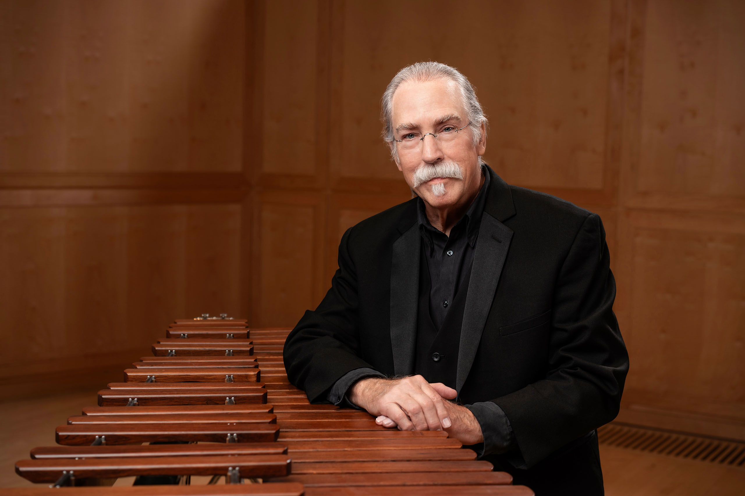 Portrait of Douglas Wolf wearing a black tuxedo, with his hands resting on a marimba, and wood paneled walls in the background.