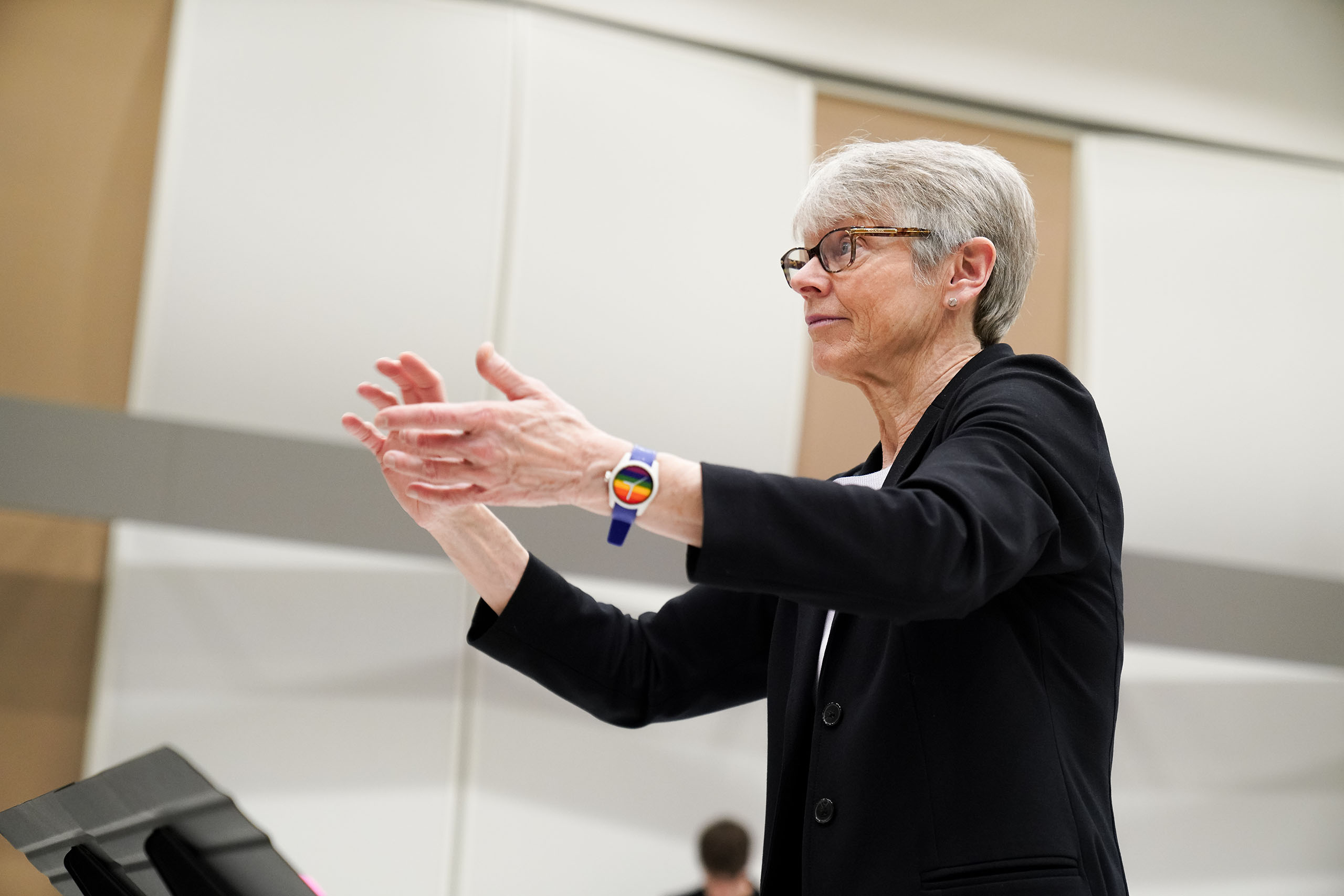In a studio space with light walls, Ellen Rowe holds up her arms to conduct, with a stand partly visible in the foreground.