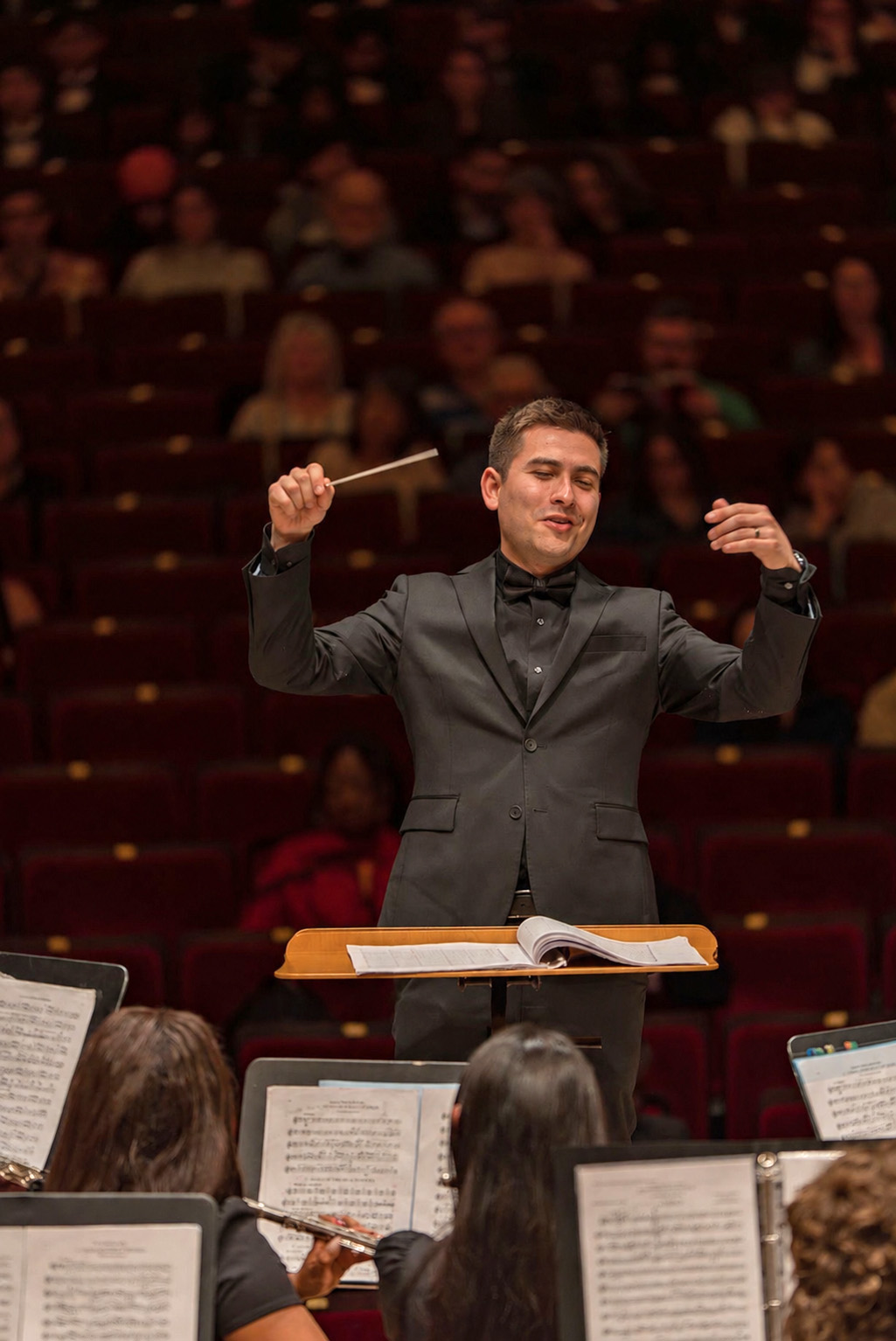 Elliot Polot conducts holding a baton in black concert attire. A darkened house of people fills the background, with the heads of flute players and music on stands in the foreground.