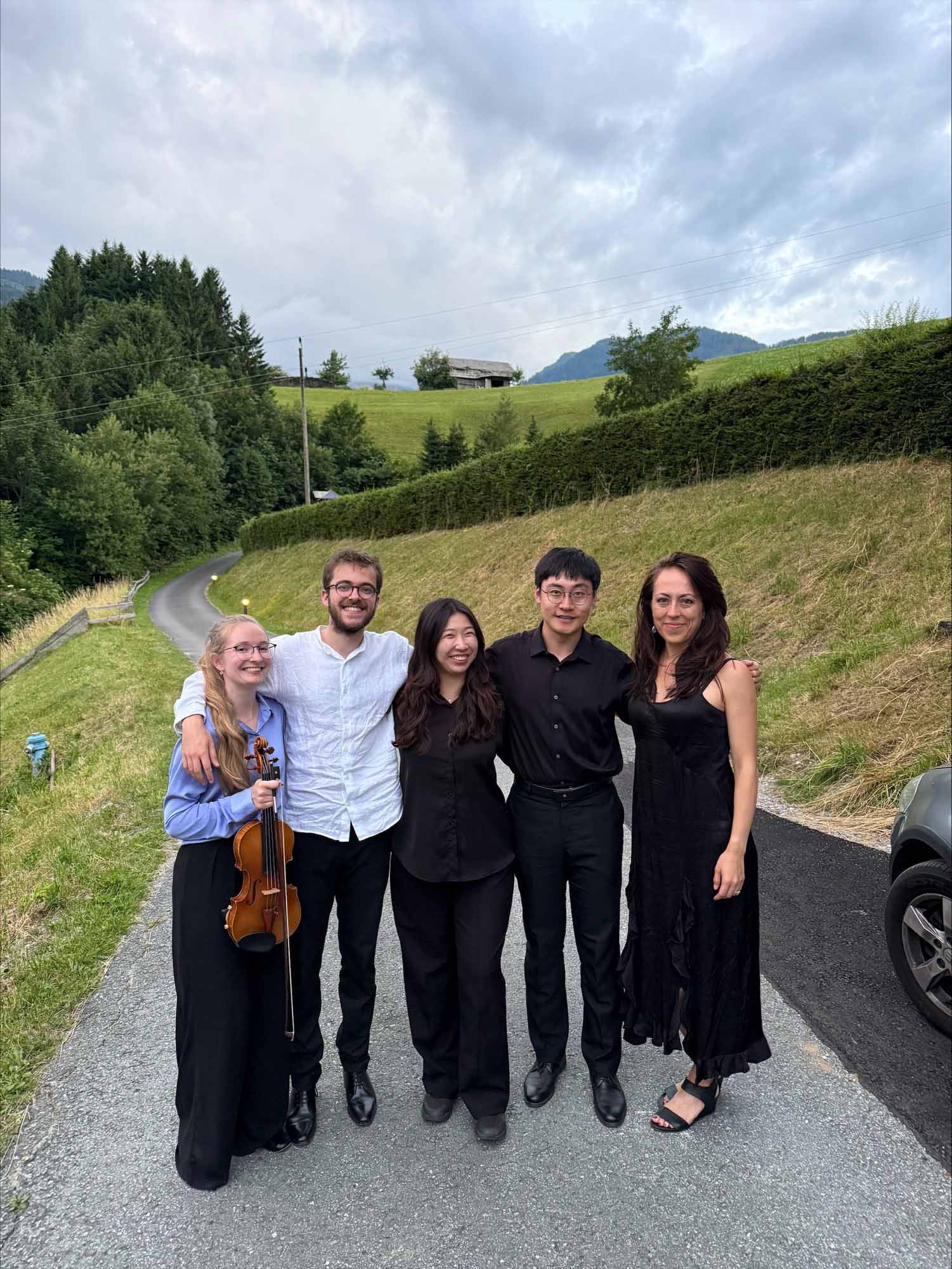 Five people pose standing together outdoors on a small country lane, with a grassy hillside and mountaintops in the background. All are wearing concert attire and one holds a viola and bow.