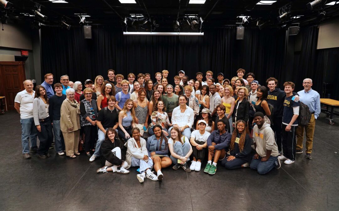 About 70 people pose together gathered closely in rows, with some seated or kneeling, in a studio space with a black curtain in the background. Ben Ahlers is in the middle of the group seated in a chair.