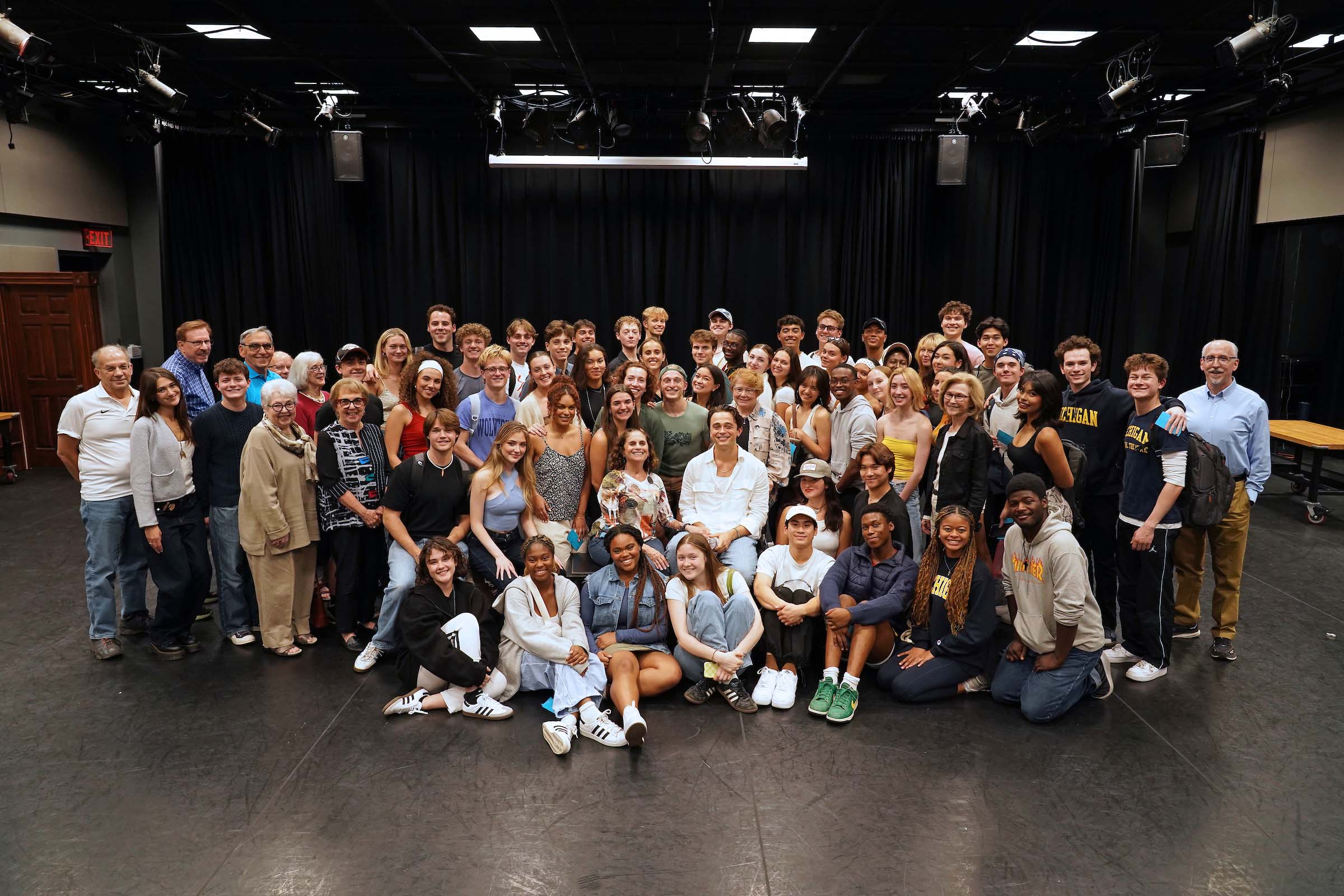 About 70 people pose together gathered closely in rows, with some seated or kneeling, in a studio space with a black curtain in the background. Ben Ahlers is in the middle of the group seated in a chair.