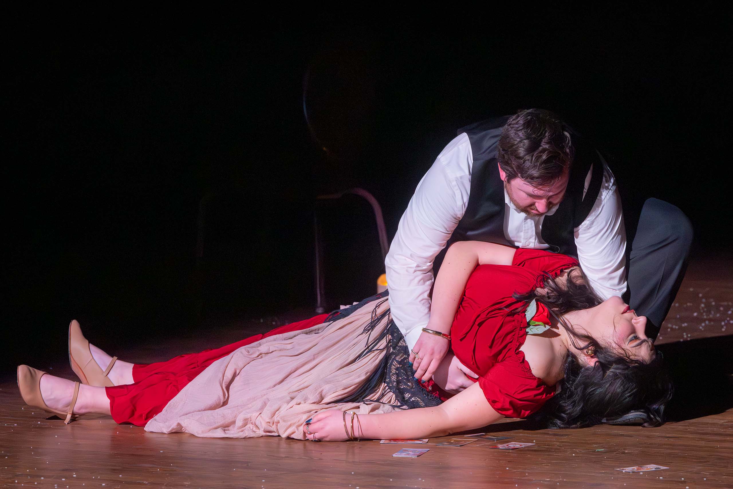 Two actors perform a death scene on stage. Carmen lies flat with limp arms and neck, costumed in a long red dress, while the other character in white shirt and black vest crouches down to hold her.