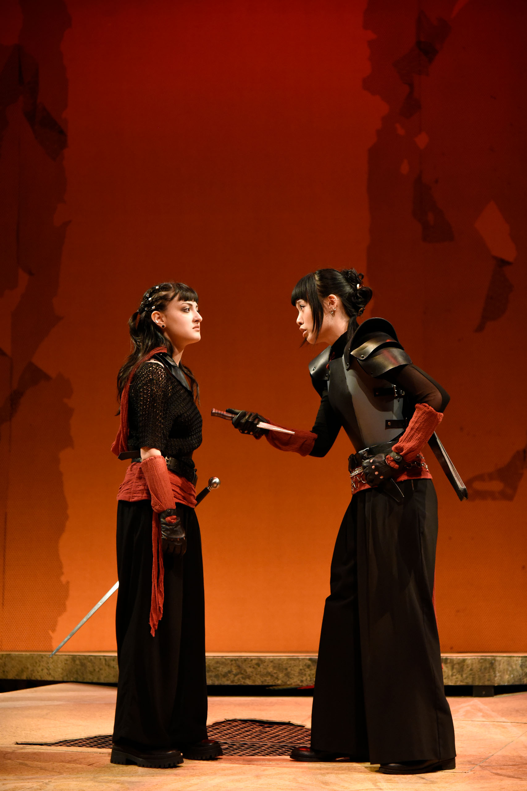 Two actors perform a tense scene standing on stage, with one holding up a knife facing inward. They wear warrior-like black costumes with red at the wrists and waist. A stage backdrop of dramatic red and orange hues rises behind them in this vertical image.
